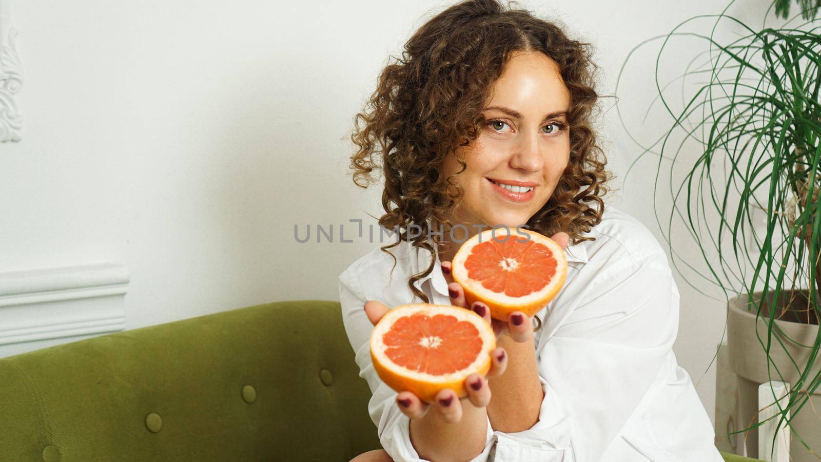 Portrait of pretty middle-aged woman with curly hair with grapefruit at home - light room. The concept of happiness, beauty and health