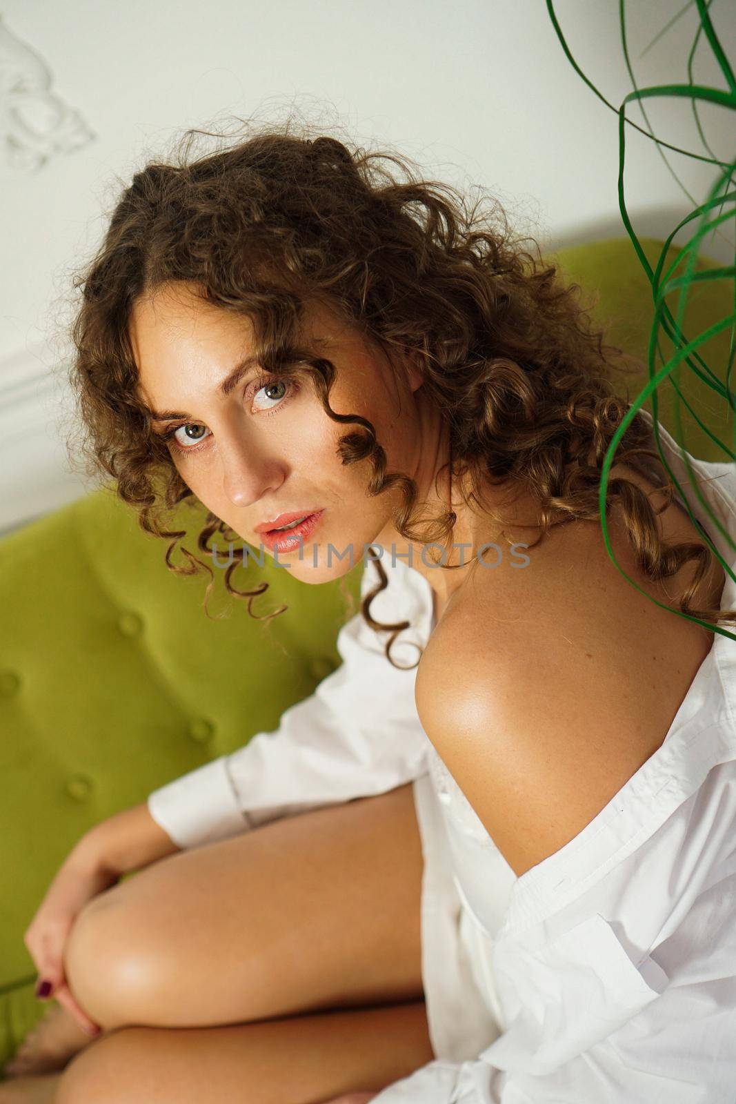 Relaxed lifestyle. Happy young woman with curly hair in white shirt spending time at home. Green sofa and white walls in room