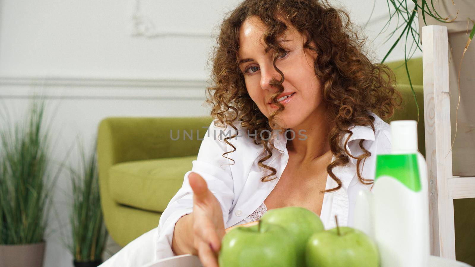 Beautiful woman with curly hair in room with green sofa. Apples and Organic Care by natali_brill