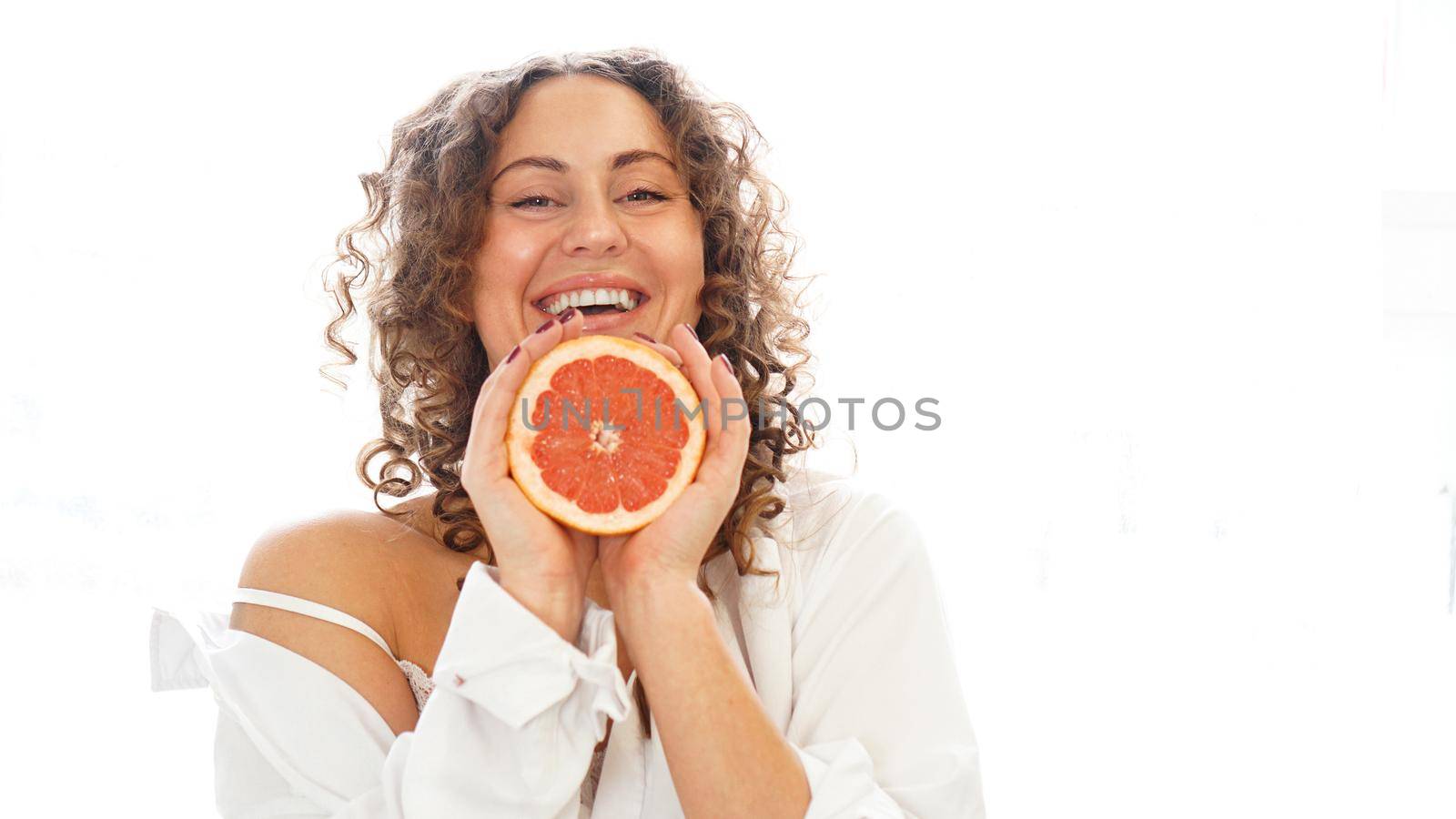 Portrait of pretty middle-aged woman with curly hair with grapefruit at home - white background. The concept of happiness, beauty and health