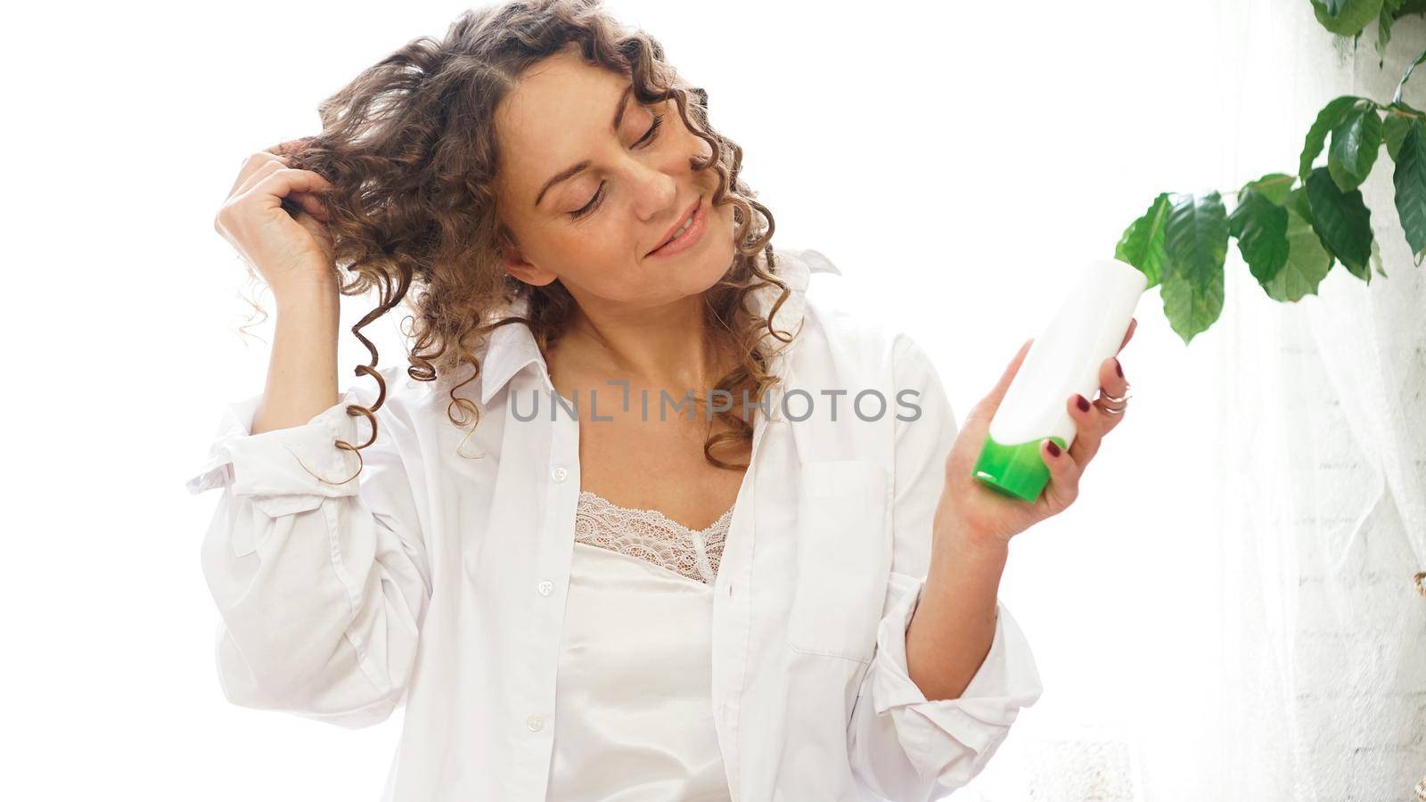 Pretty woman showing white cosmetic tube for curly hair care in her hand at home, white room with green plants