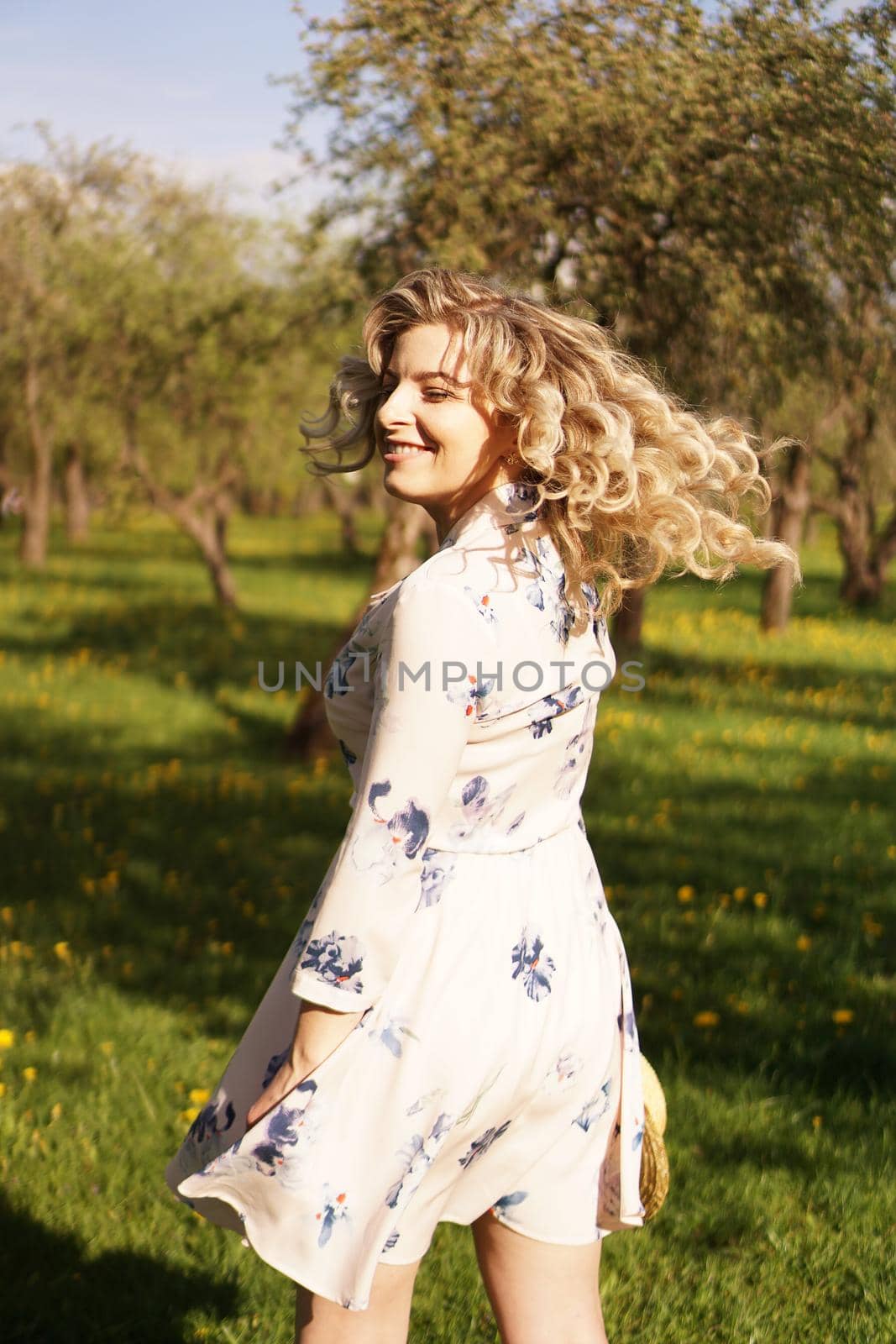 Happy young woman with blonde hair, wearing a dress, posing outdoors in a garden with cherry trees in the sun, smiling. Curls fluttering in the wind