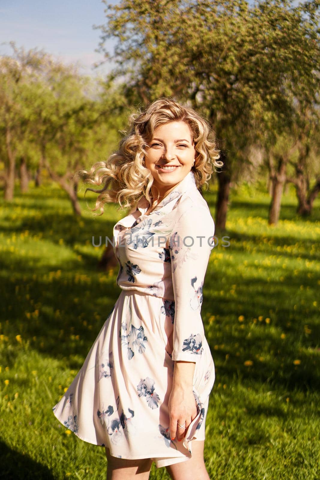 Happy young woman with blonde hair, wearing a dress, posing outdoors in a garden with cherry trees in the sun, smiling. Curls fluttering in the wind