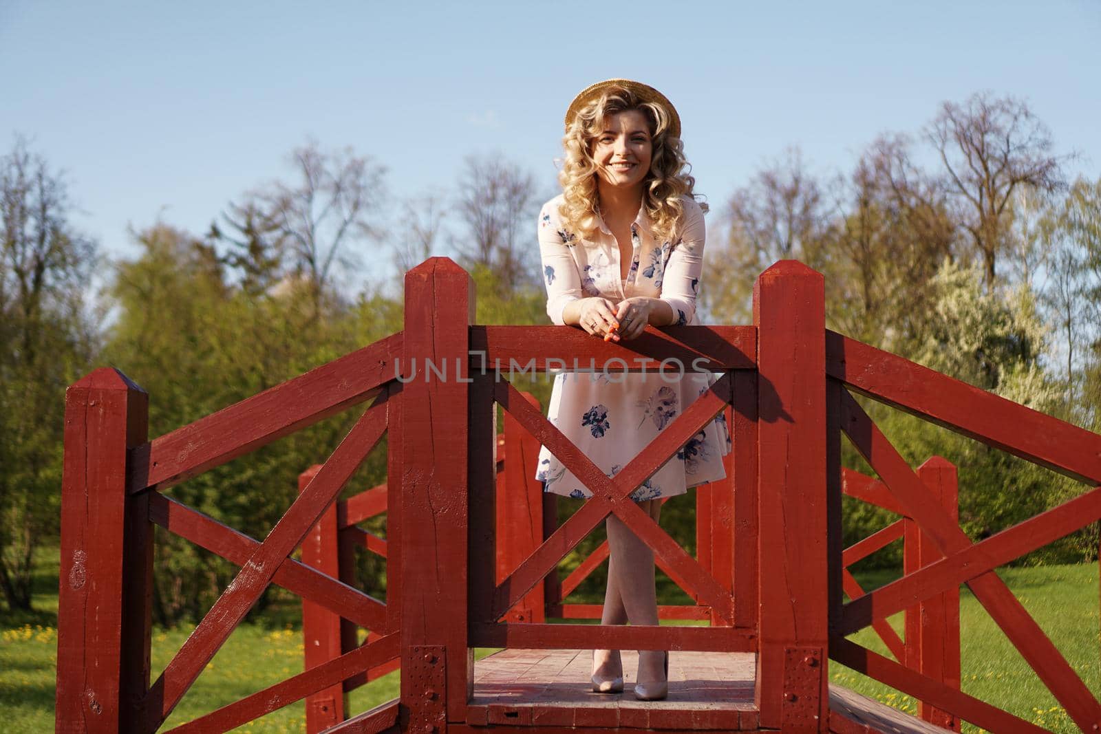 Beautiful woman in a white summer dress and a straw hat stands on a birch bridge in the park