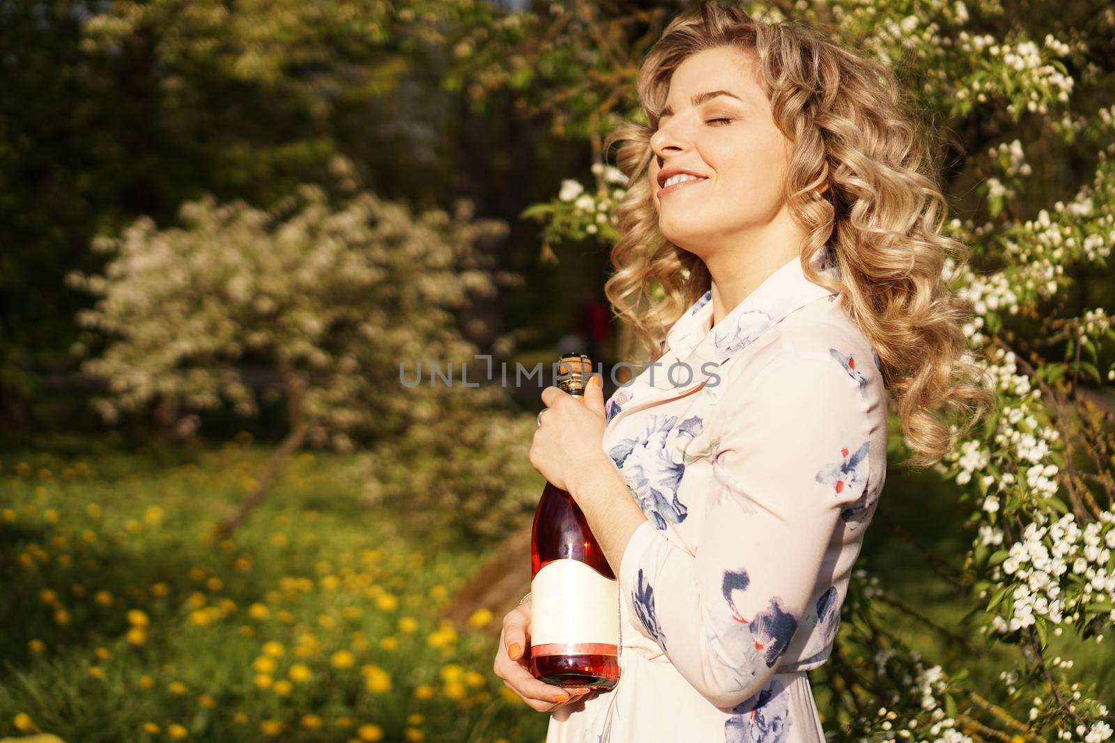 Beautiful young woman holding a blank bottle of wine while having lunch outdoors. Picnic in the park with green grass