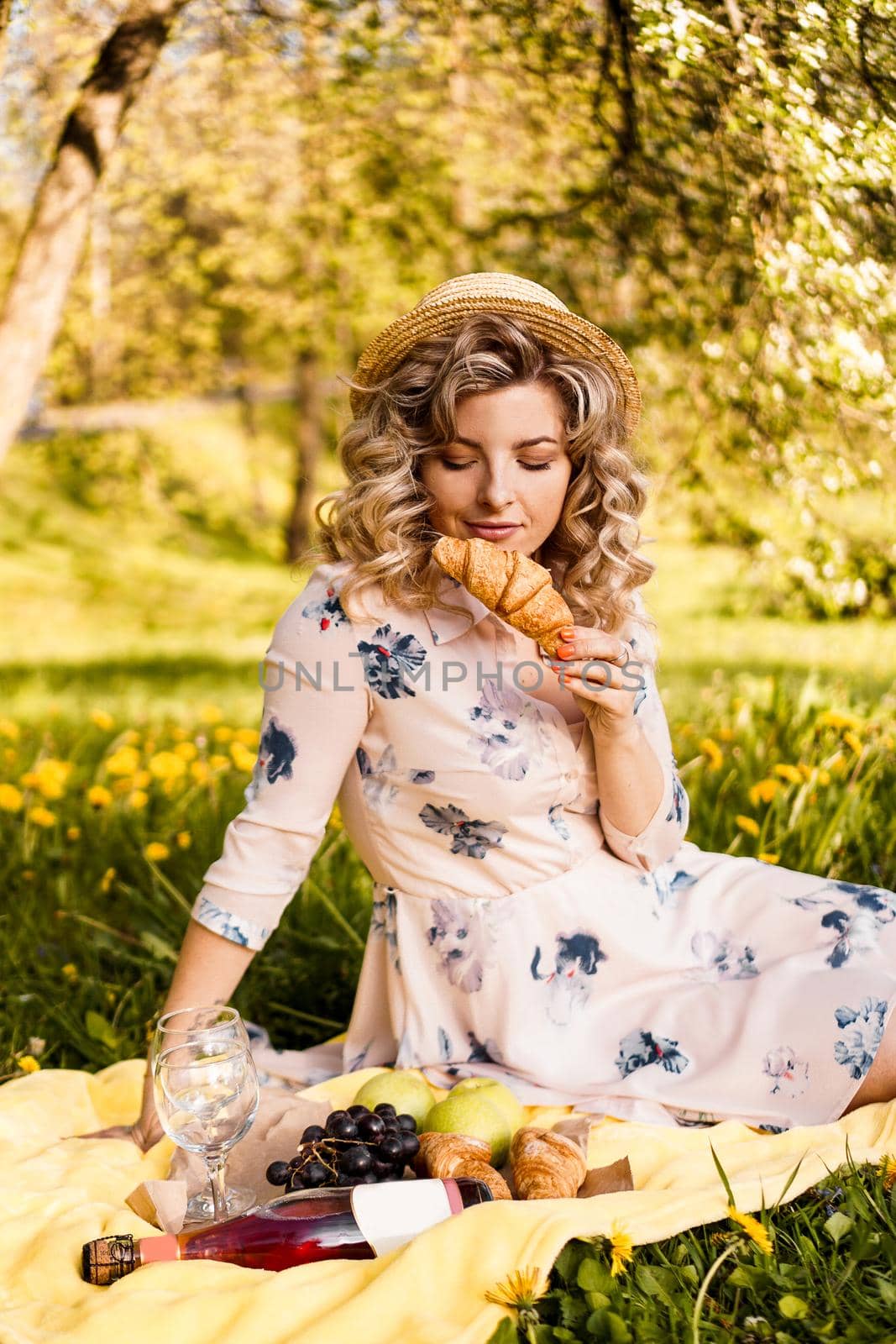 Beautiful young woman with blonde hair in straw hat and white dress lying on the plaid in the park. Woman with a croissant on a picnic