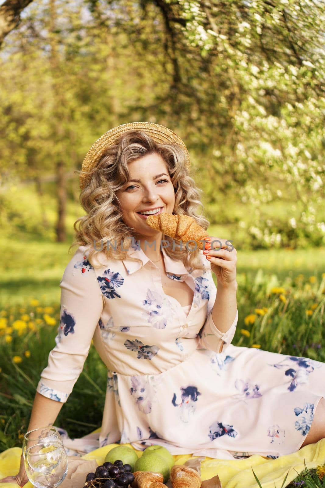 Beautiful young woman with blonde hair in straw hat and white dress lying on the plaid in the park. Woman with a croissant on a picnic