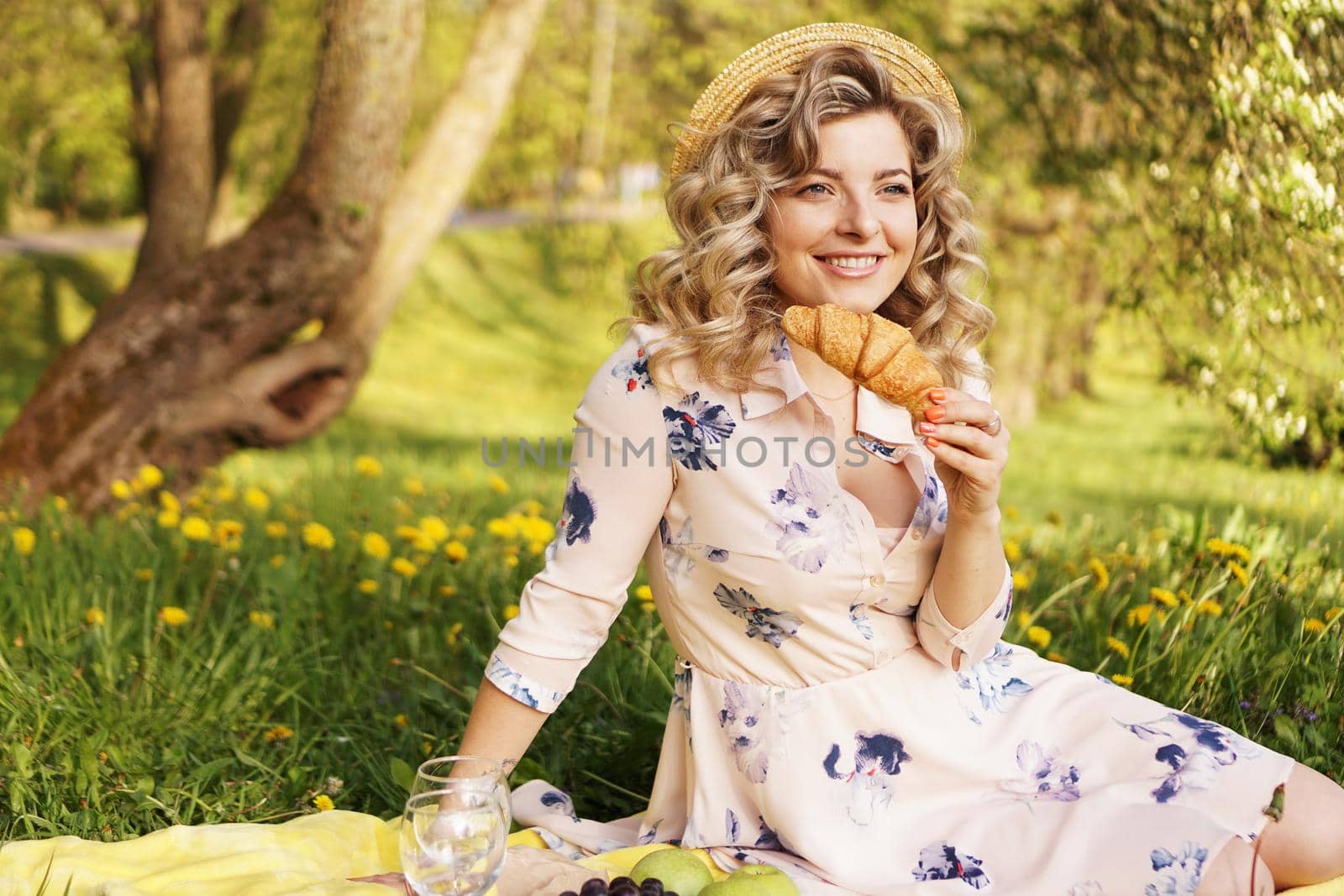 Beautiful young woman with blonde hair in straw hat and white dress lying on the plaid in the park. Woman with a croissant on a picnic