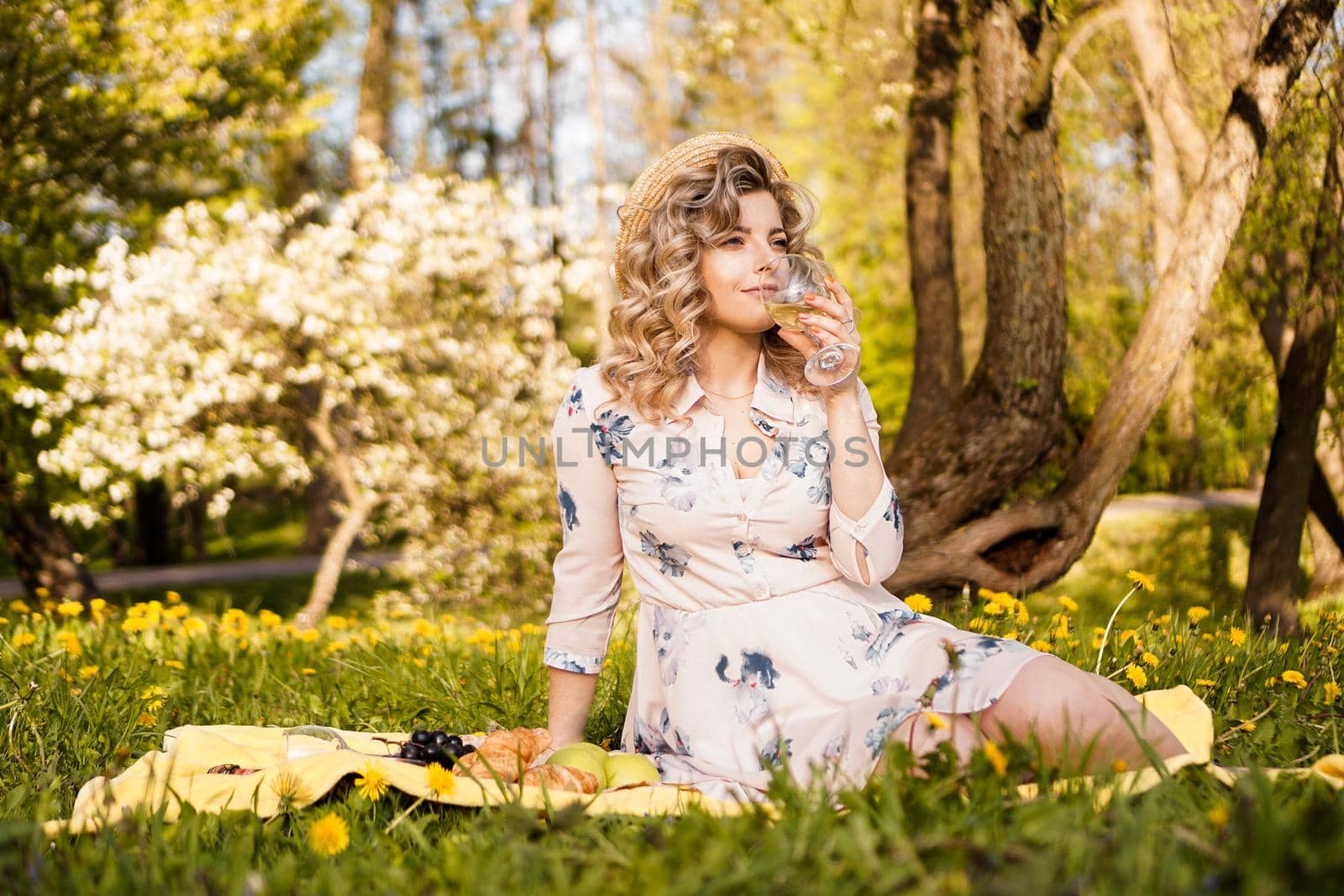 Beautiful young woman with blonde hair in straw hat drinks wine and sitting on the plaid in the garden during a summer picnic
