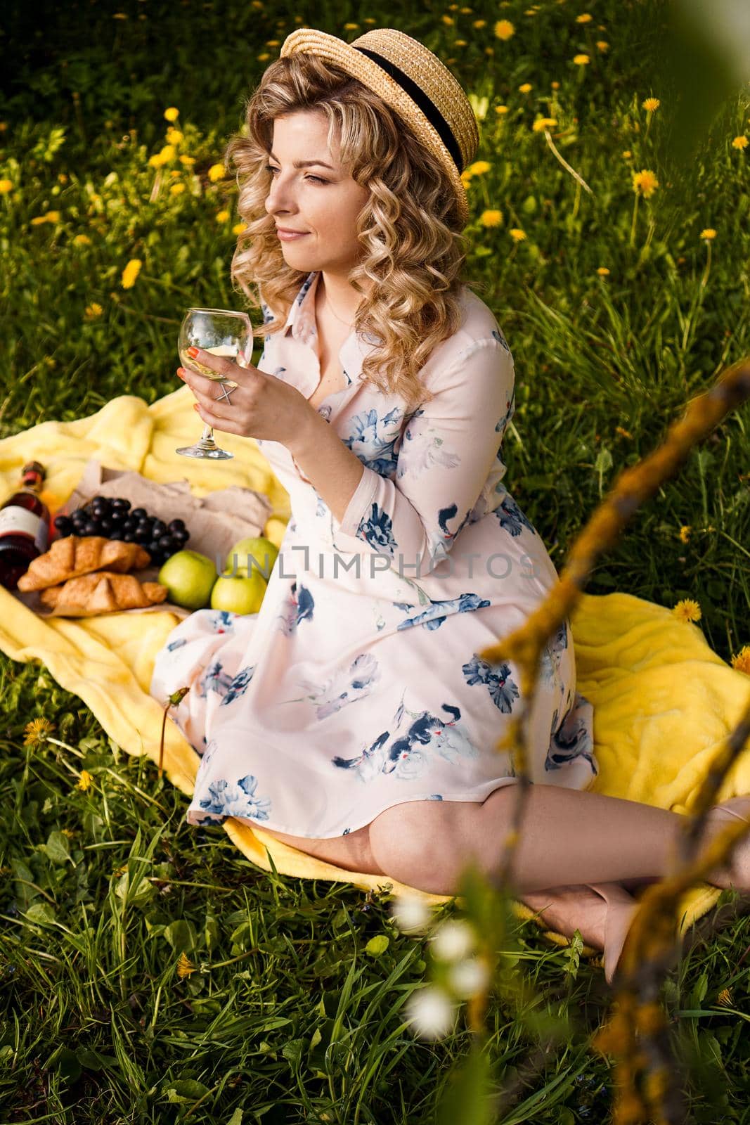 Beautiful young woman with blonde hair in straw hat drinks wine and sitting on the plaid in the garden during a summer picnic