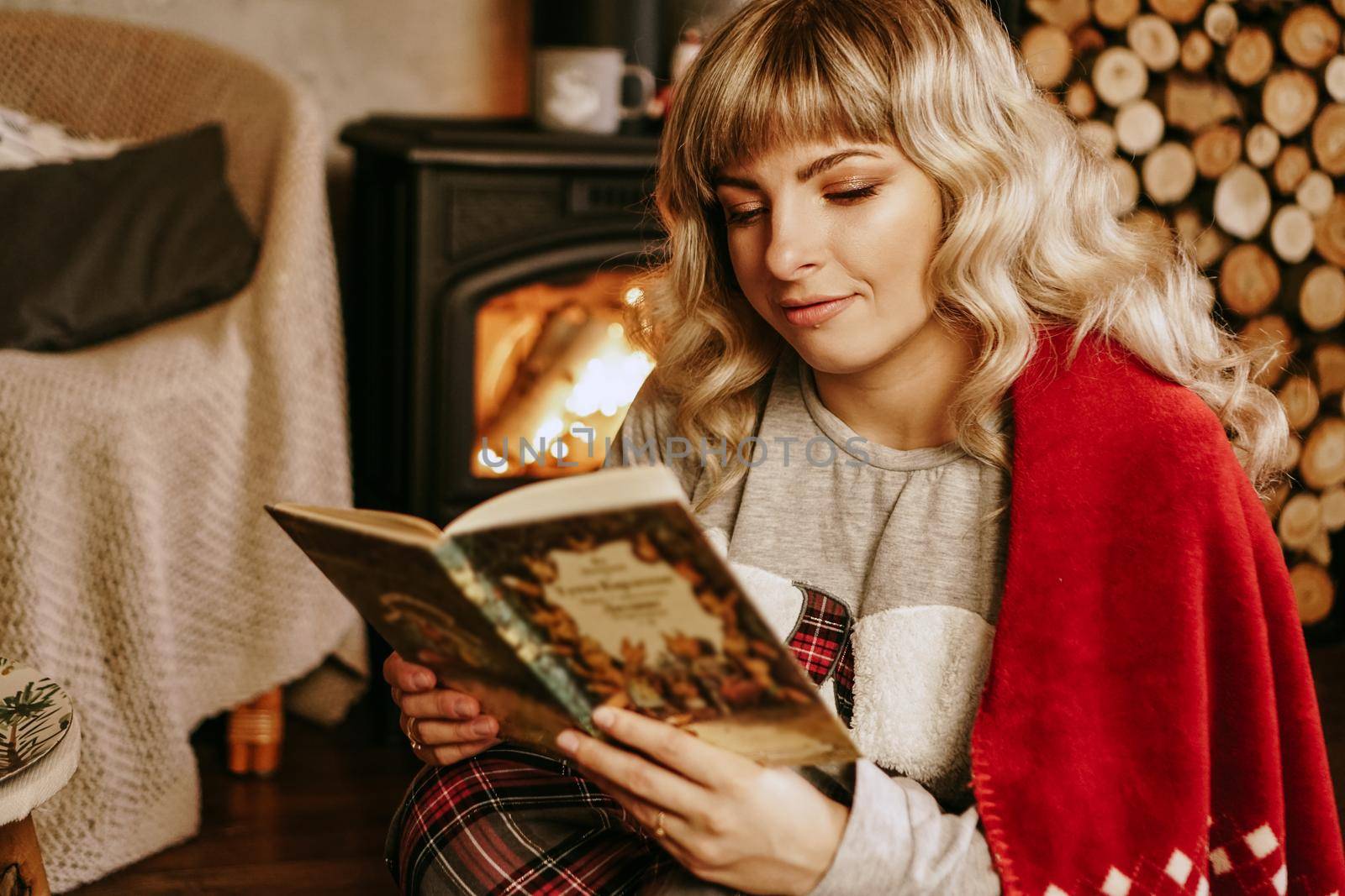 Happy young woman reading book in front of christmas interior with fireplace. The girl is dressed in homely cozy clothes with a red warm plaid