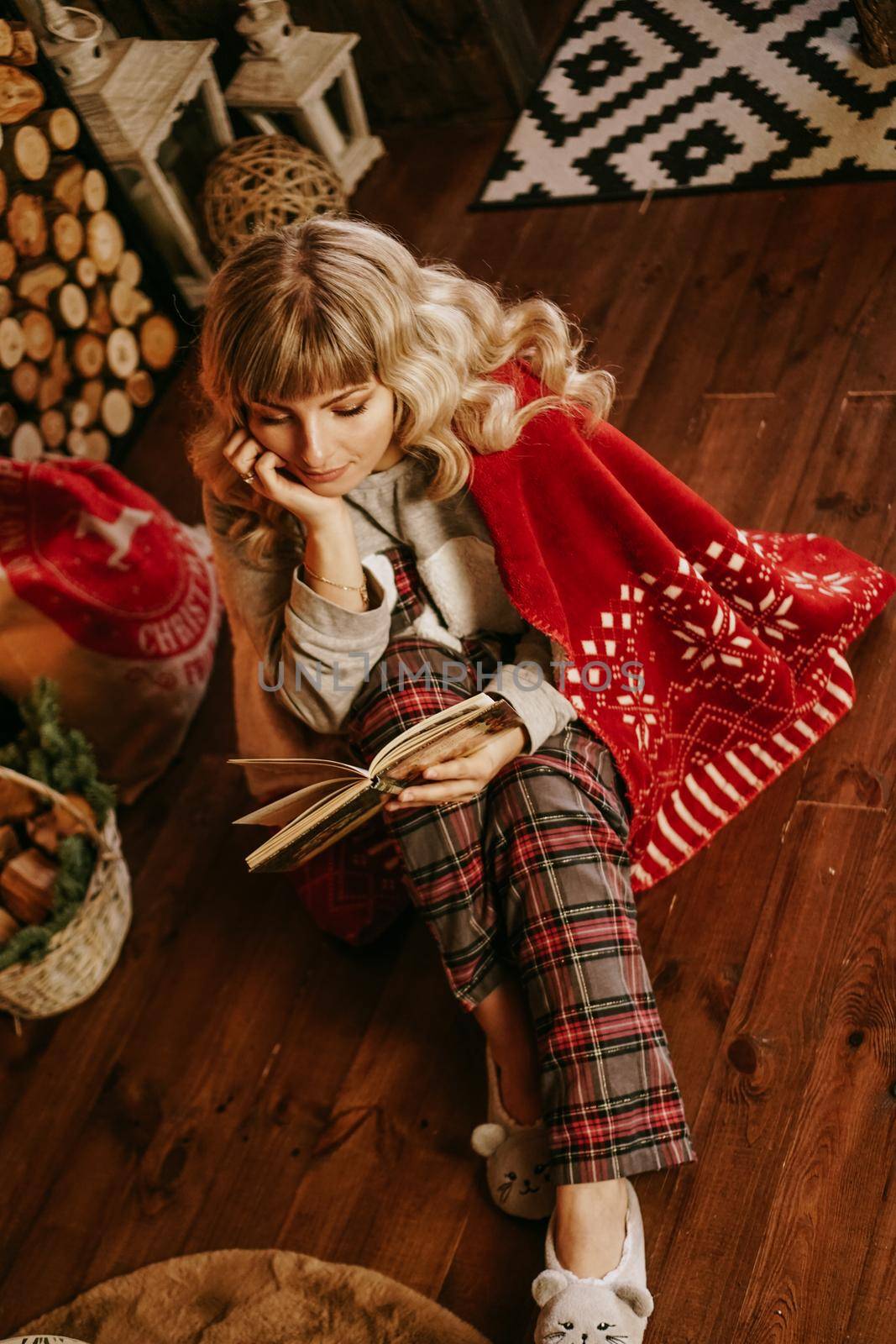 Happy young woman reading book in front of christmas interior with fireplace. The girl is dressed in homely cozy clothes with a red warm plaid