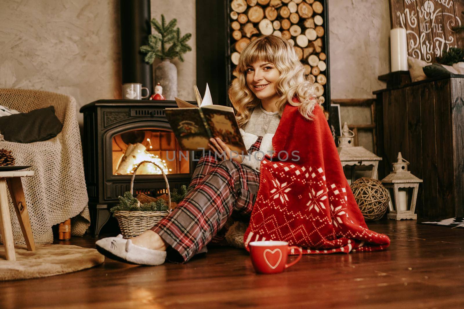 Happy young woman reading book in front of christmas interior with fireplace. The girl is dressed in homely cozy clothes with a red warm plaid