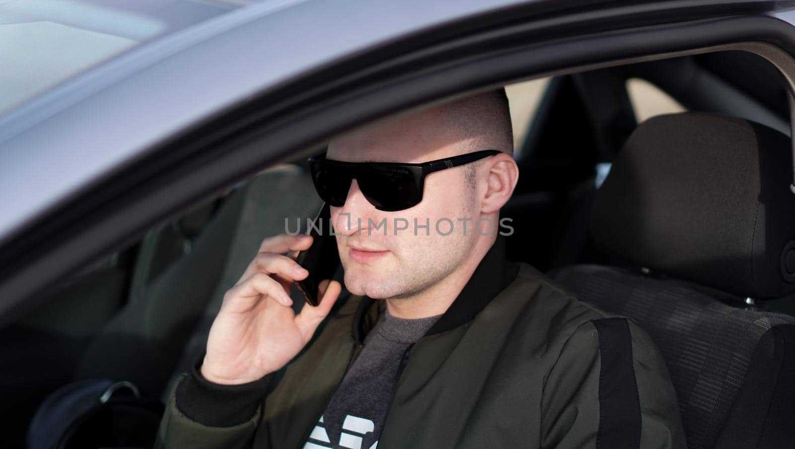 Man talking on a mobile phone while driving car. Side view of confident young stylish man in sunglasses driving his car