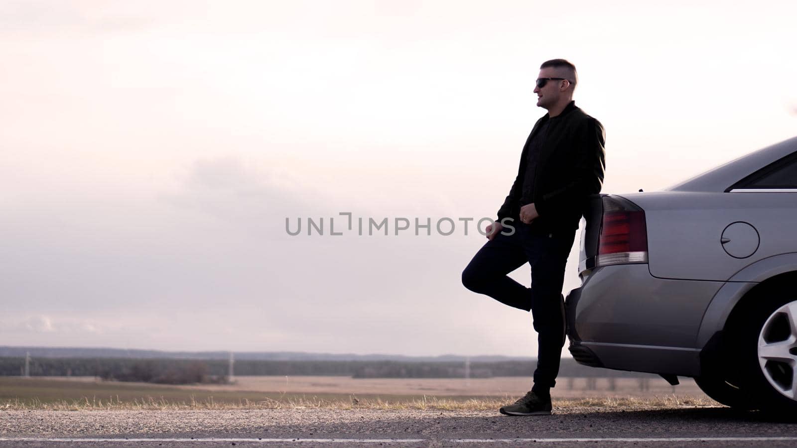 Sde view of young businessman in eyeglasses sitting on car trunk at nature background
