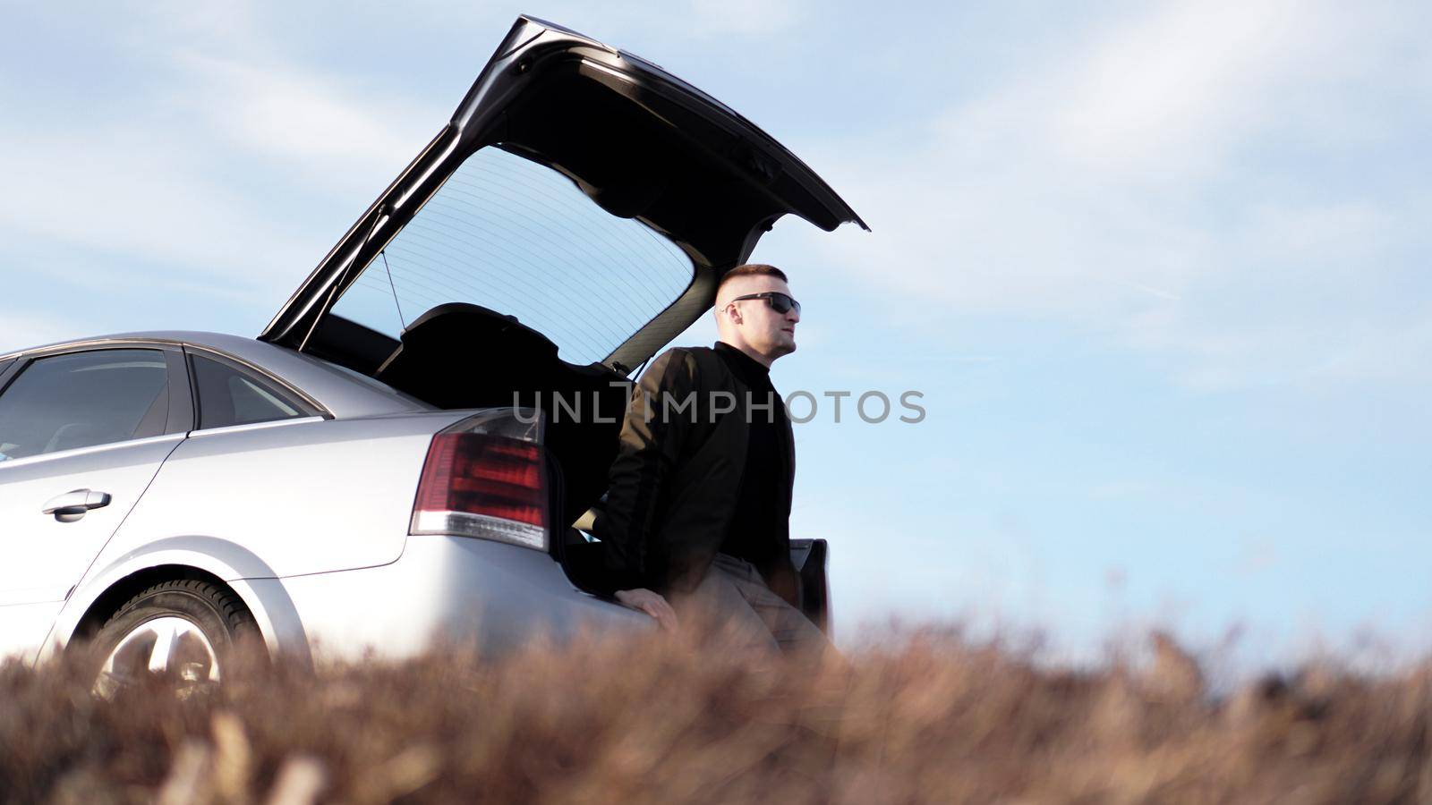 Side view of young businessman in eyeglasses sitting on car trunk at nature background