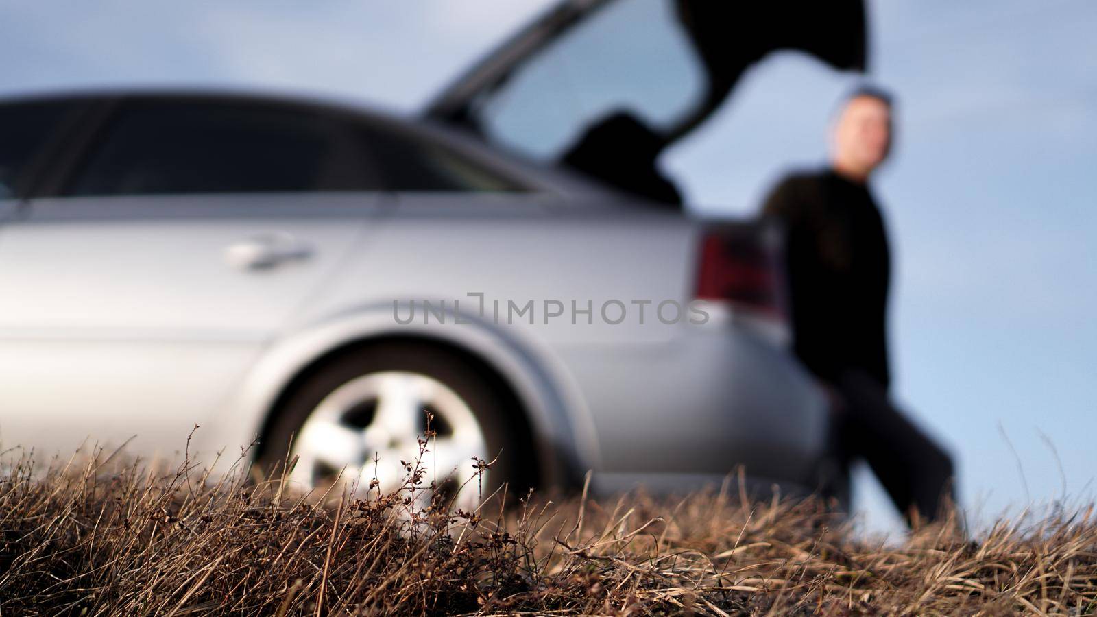 Side view of young businessman in eyeglasses sitting on car trunk at nature - blurred background for banner