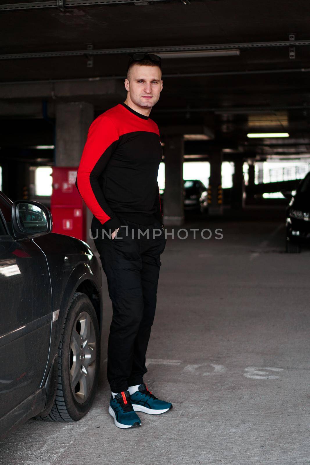 A young man in a tracksuit stands next to a car in an underground parking lot. Vertical photo