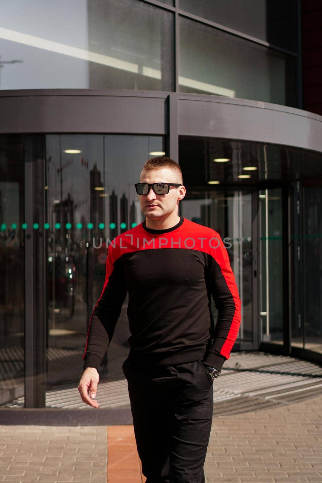 A young athletic man in sunglasses in a tracksuit walks out of the revolving doors of a hotel or shopping mall