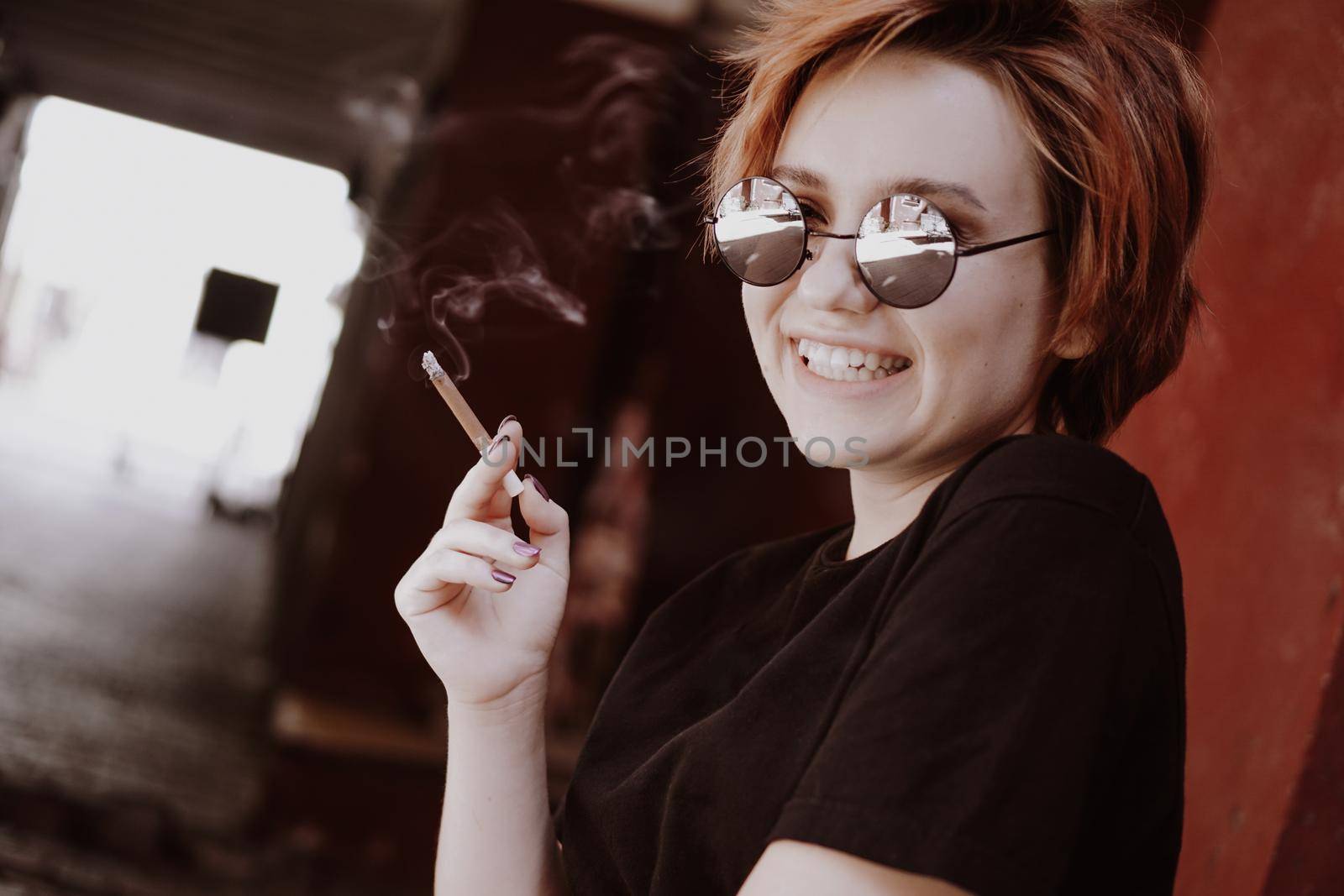 Millennial cool pretty girl with short red hair and mirror sunglasses smoking cigarette in the old city with red walls. Smiling happy teen girl