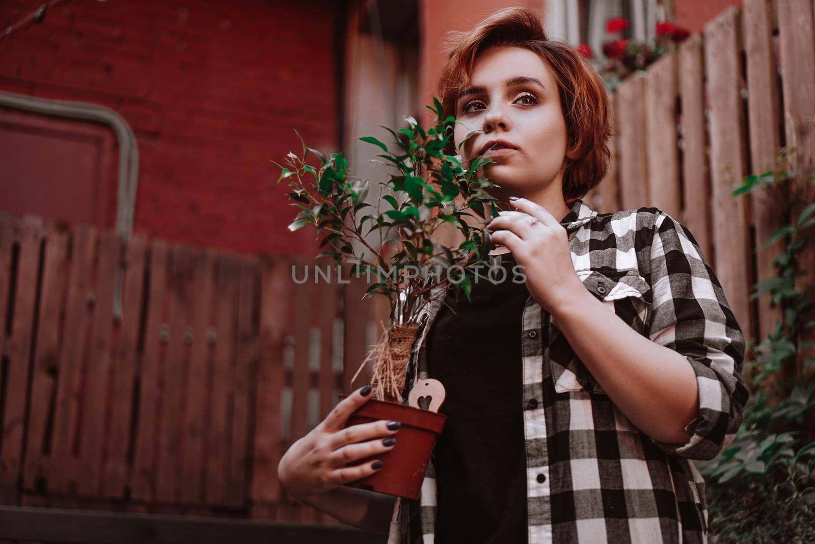 Beautiful young woman with short red hair in a plaid shirt holding a flower in a pot in the backyard