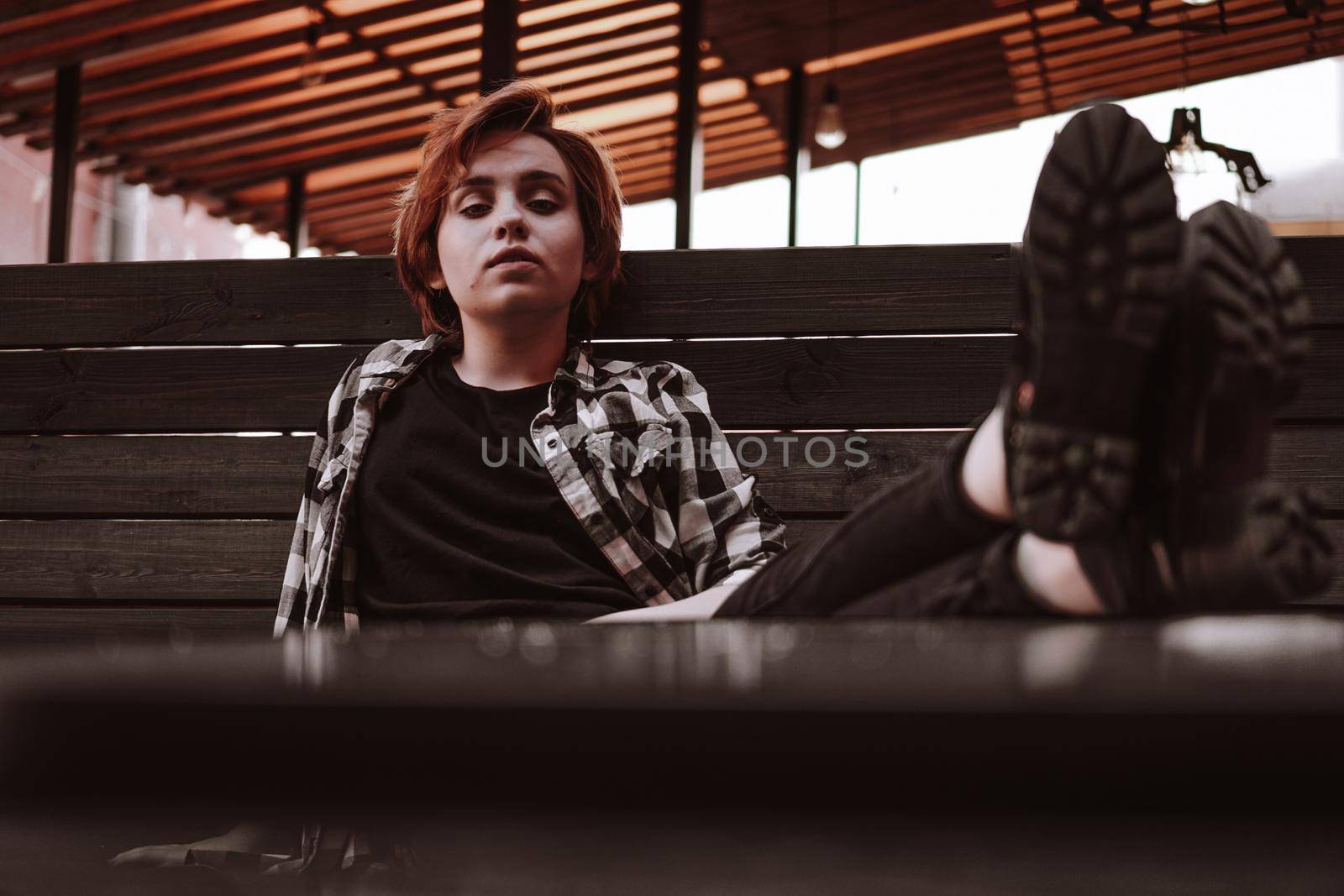 Young woman with short red hair in a plaid shirt in a bar put her legs on table