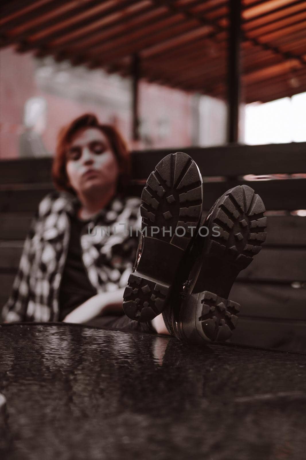 Young woman with short red hair in a plaid shirt in a bar put her legs on table