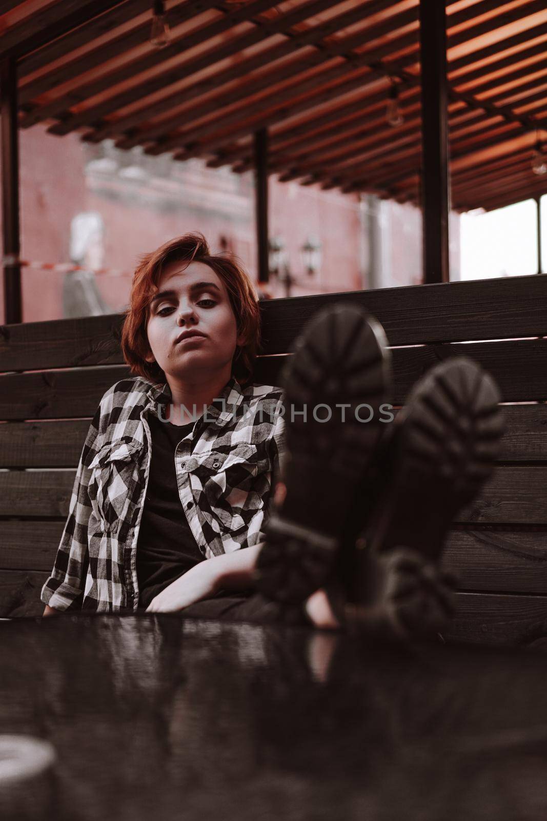 Young woman with short red hair in a plaid shirt in a bar put her legs on table