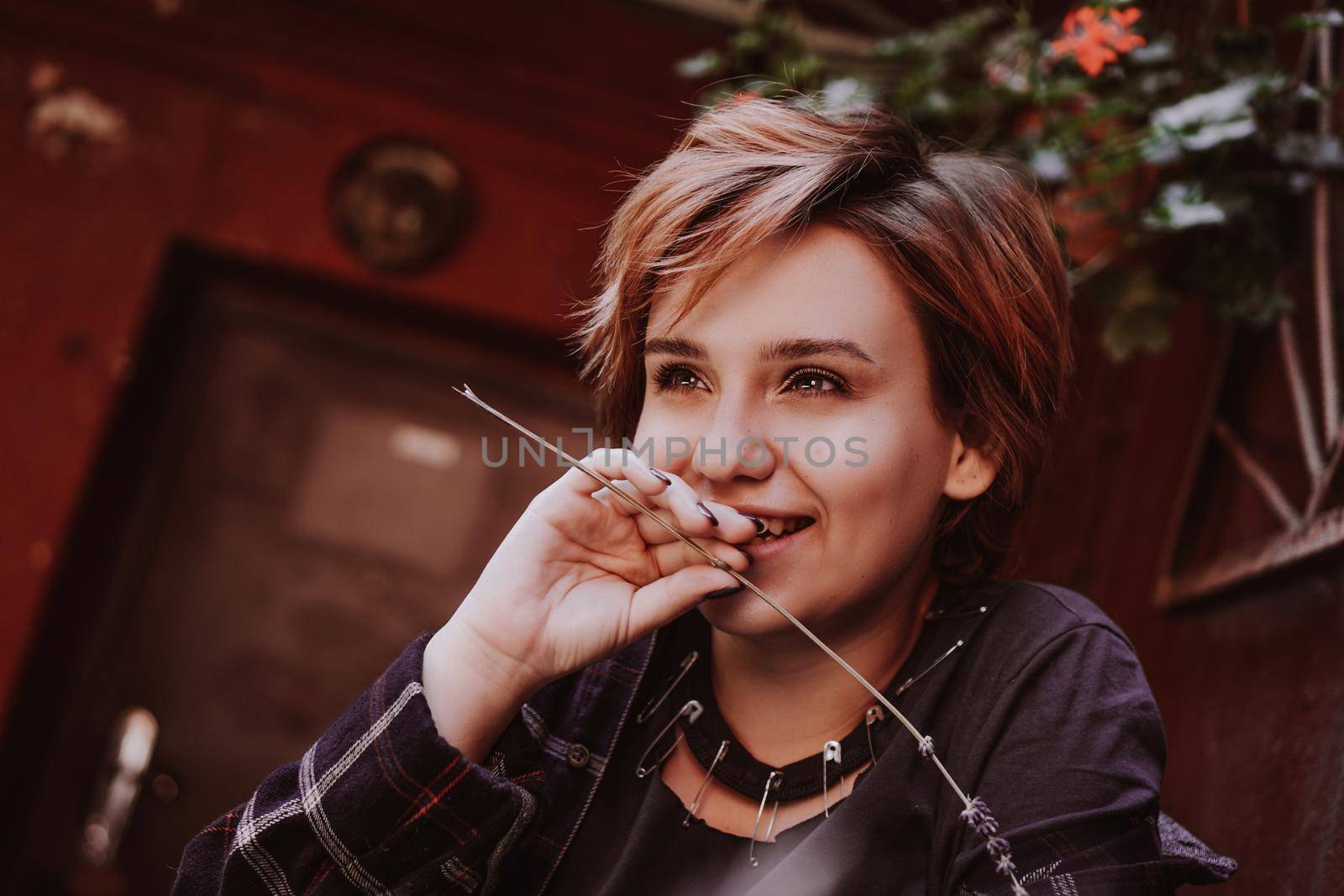 Outdoor portrait of young beautiful woman with short red hair posing with lavender flower. City lifestyle. Girl in the old city with red walls