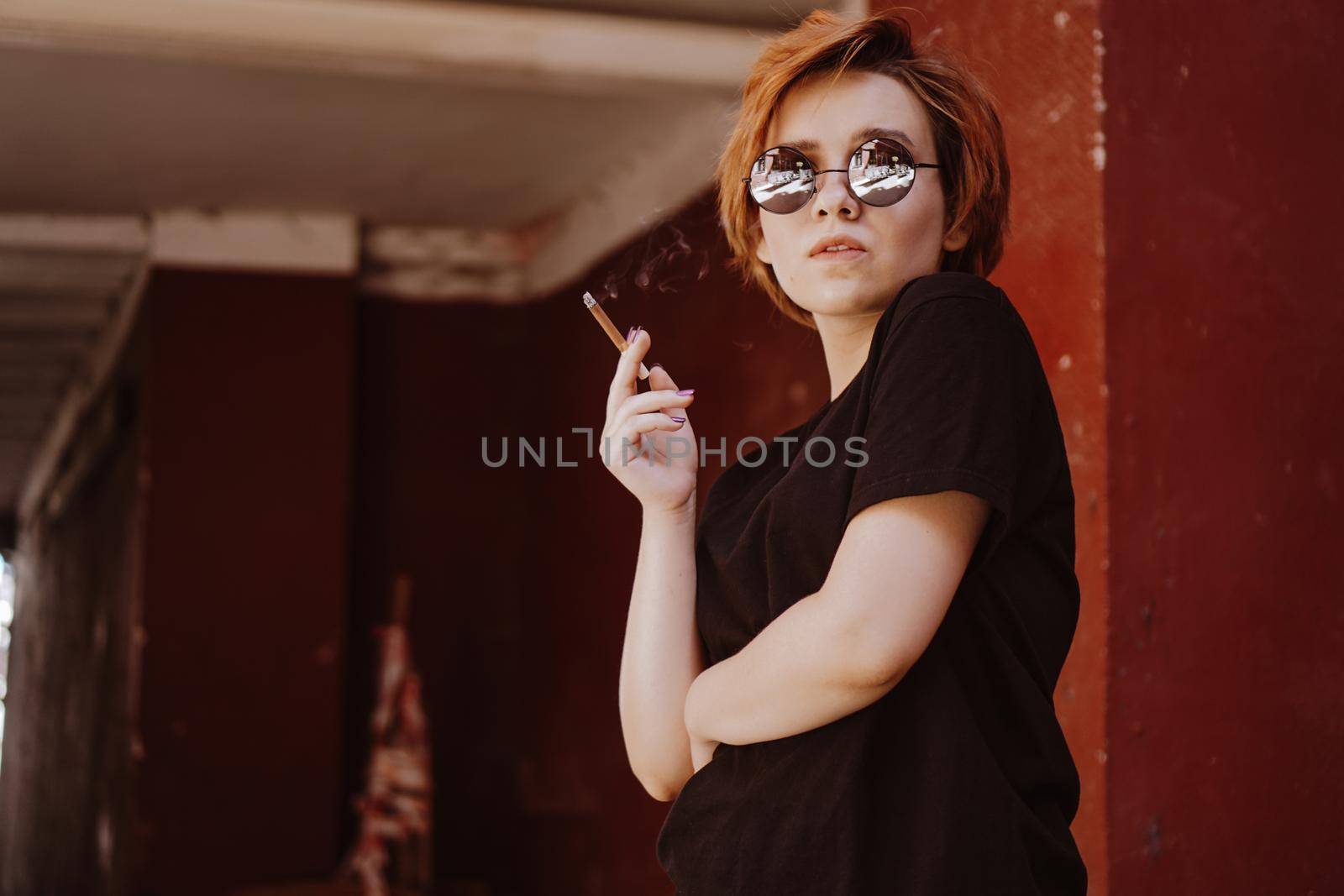 Millennial cool pretty girl with short red hair and mirror sunglasses smoking cigarette in the old city with red walls