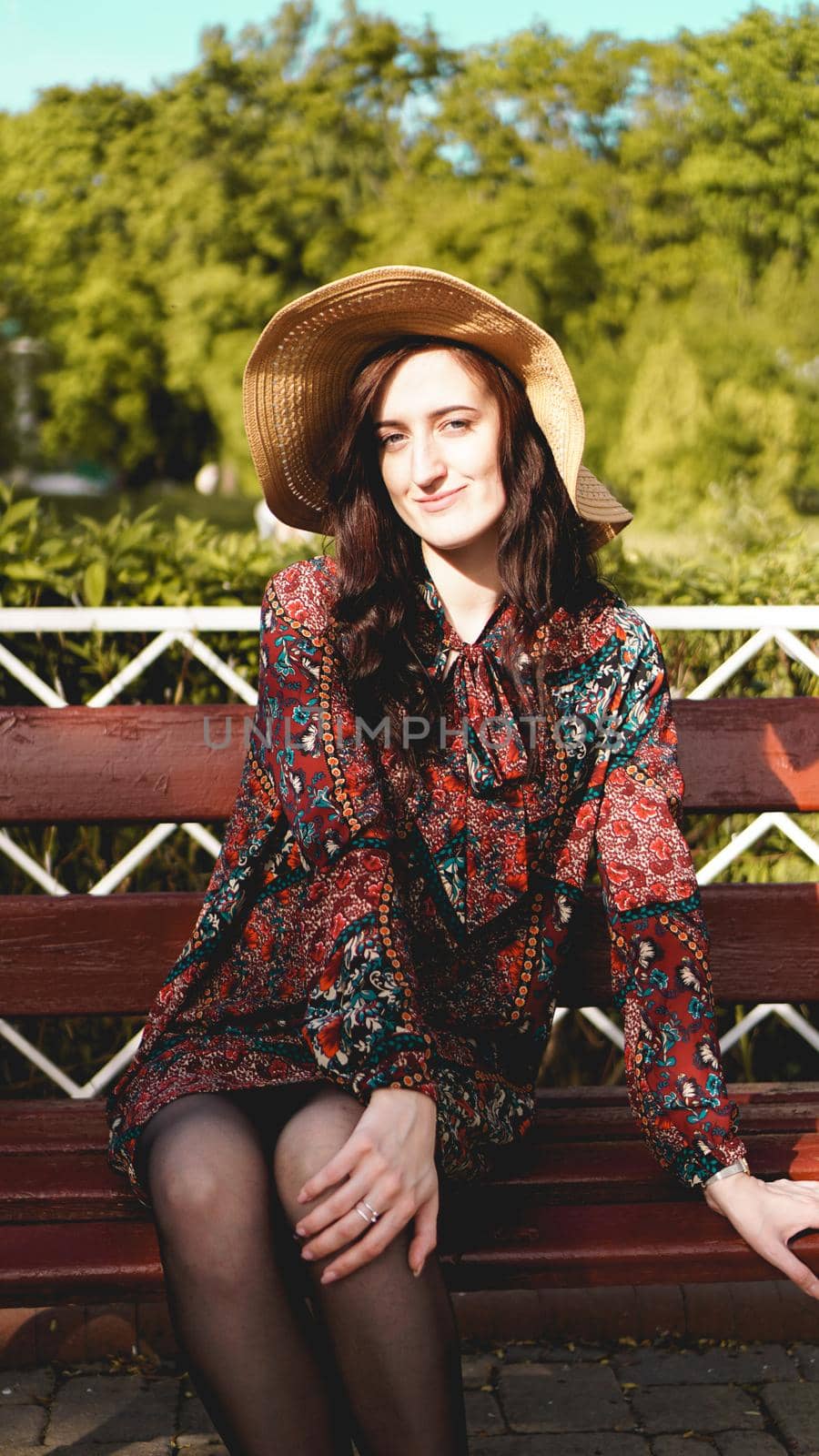 Fashionably dressed young pretty woman on the street on a sunny day. Beautiful girl in dress and hat is sitting on a bench and smiling. Street-fashion concept