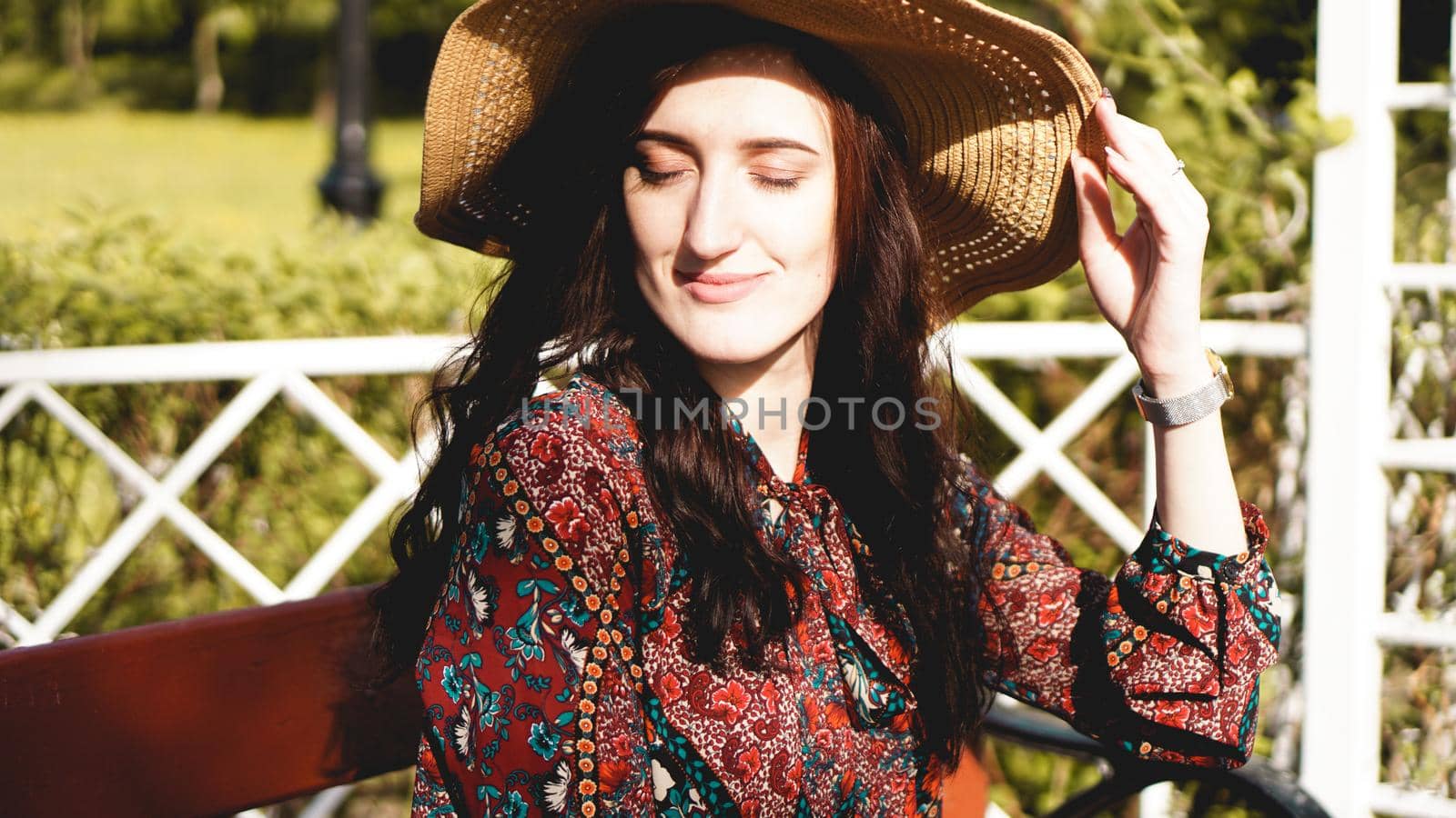 Fashionably dressed young pretty woman on the street on a sunny day. Beautiful girl in dress and hat is sitting on a bench and smiling. Street-fashion concept