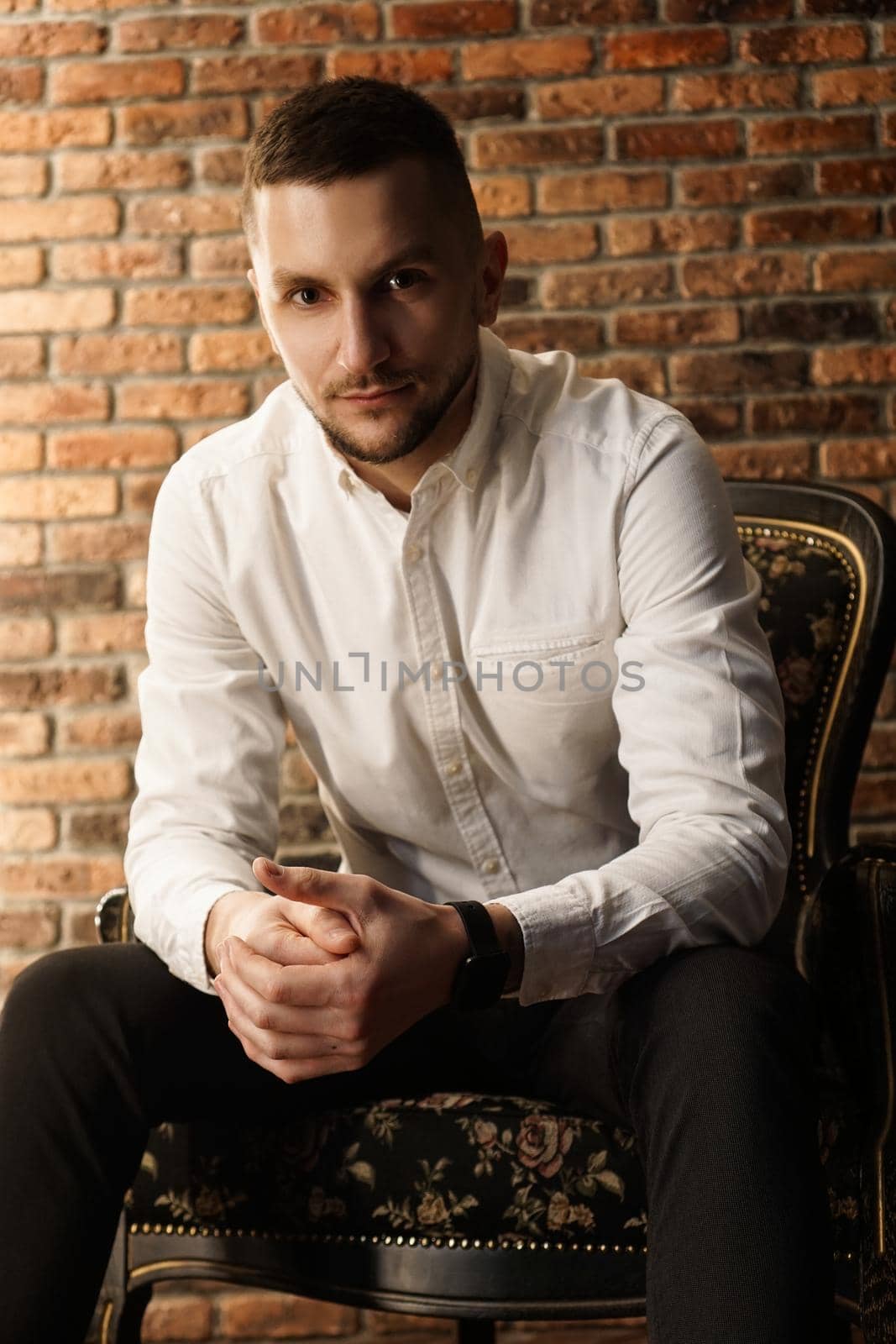 Portrait handsome fashionable man in a white shirt sits on a chair in a photo studio loft. Young businessman is thinking about future