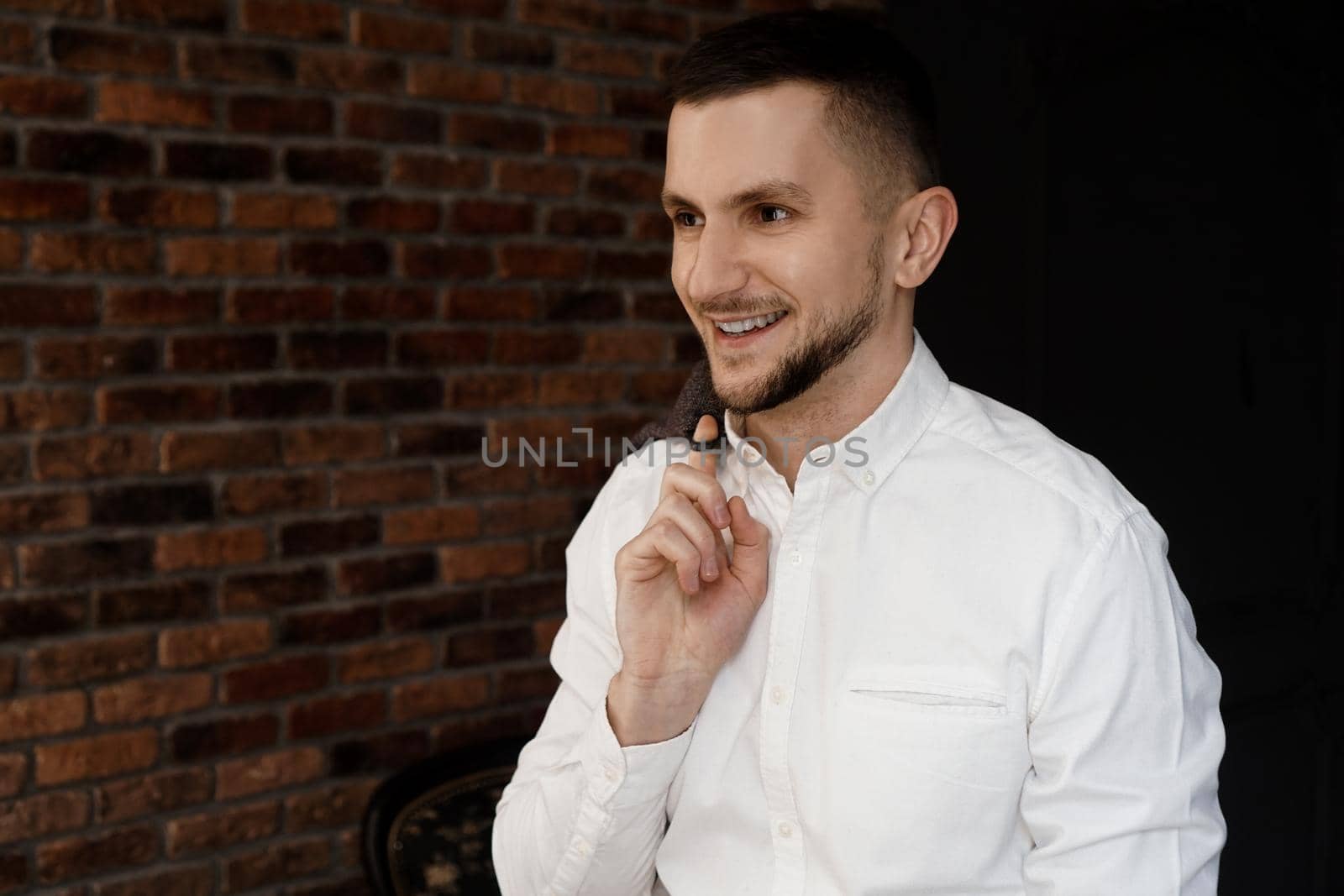 Stylish young businessman in white shirt is smiling and holding a jacket on finger, standing against brick wall