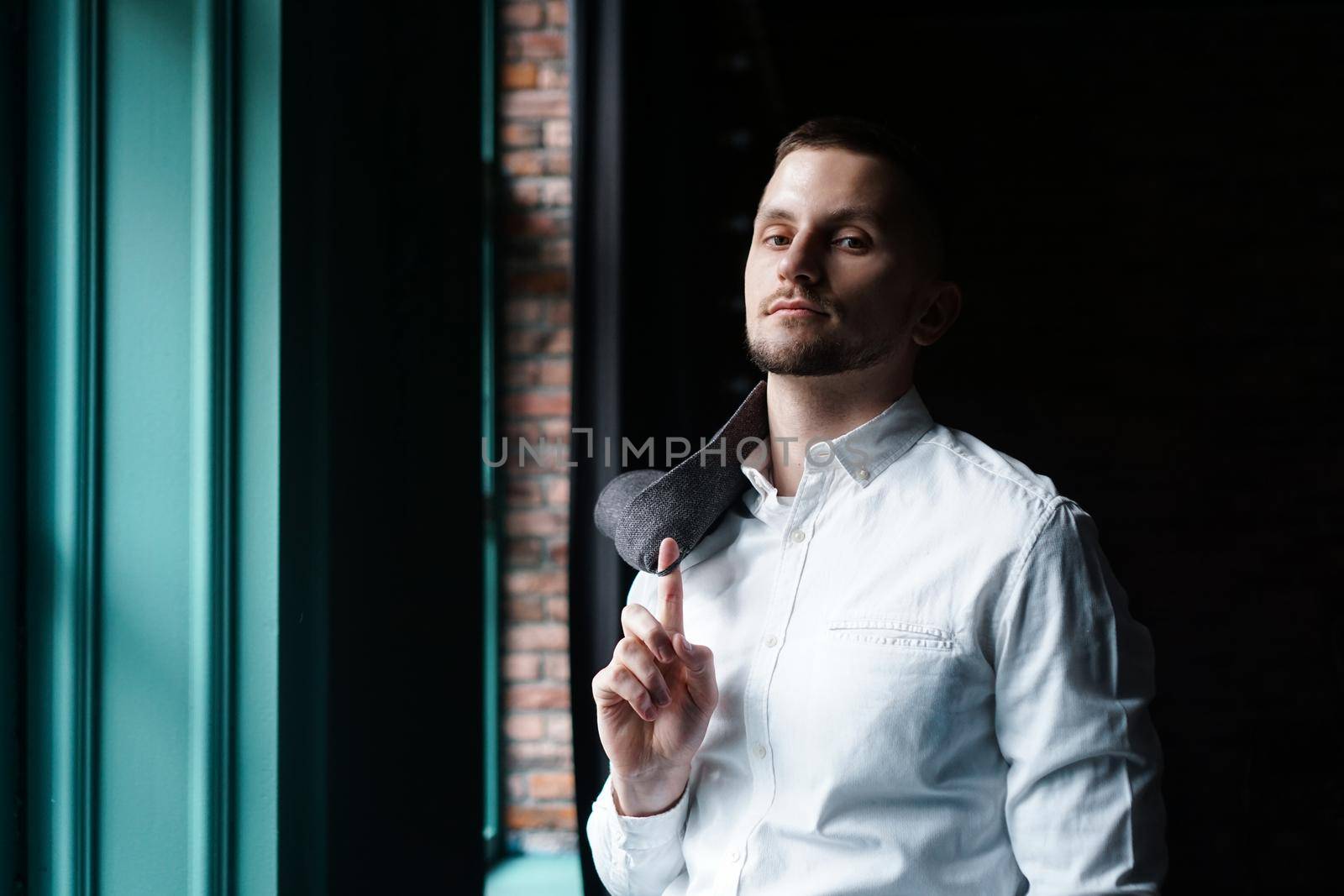 View in profile of a young businessman, dressed in a white shirt standing near the window on a dark walls and looking at the camera