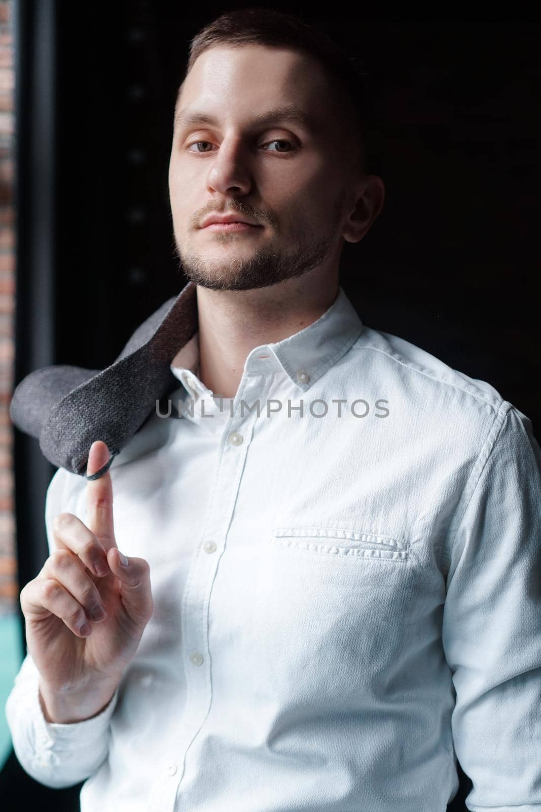 Stylish young businessman in white shirt is holding a jacket on finger, standing against black wall
