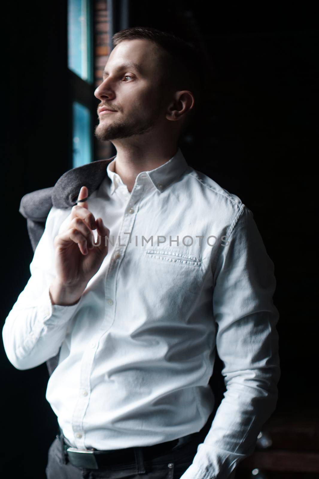 View in profile of a young businessman, dressed in a white shirt standing near the window on a dark walls and looking out the window. Vertical photo