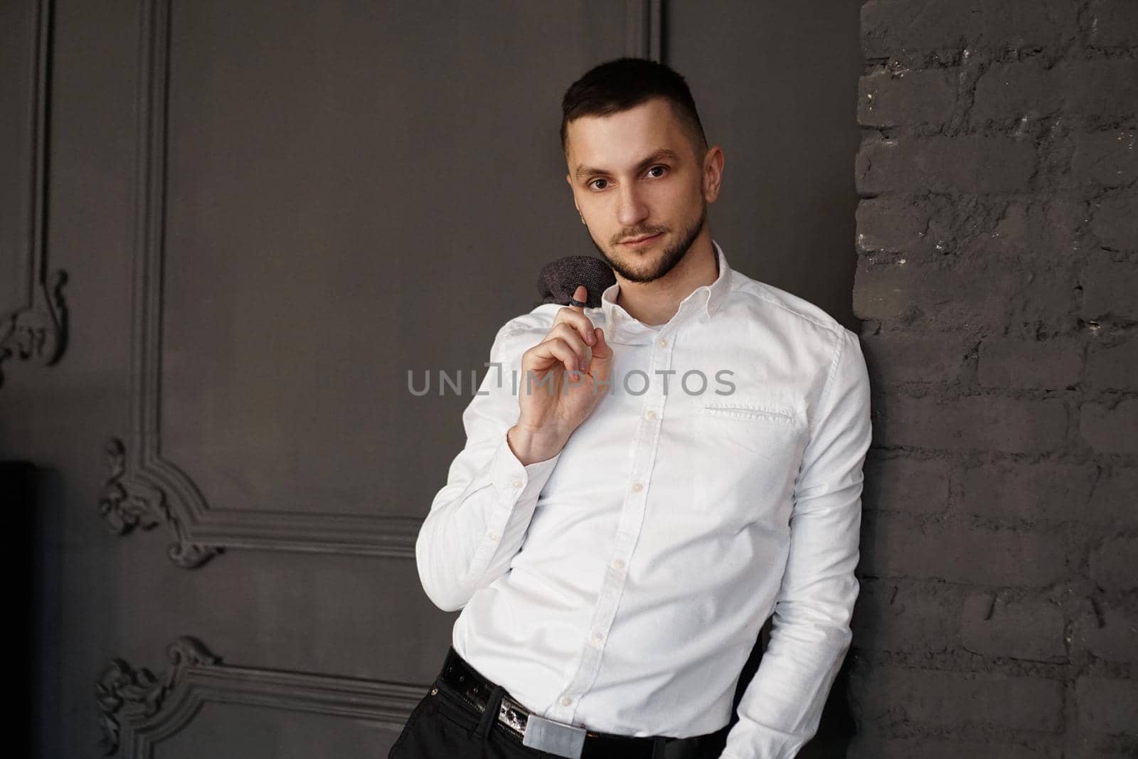 Stylish young businessman in white shirt is holding a jacket on finger, standing against brick wall