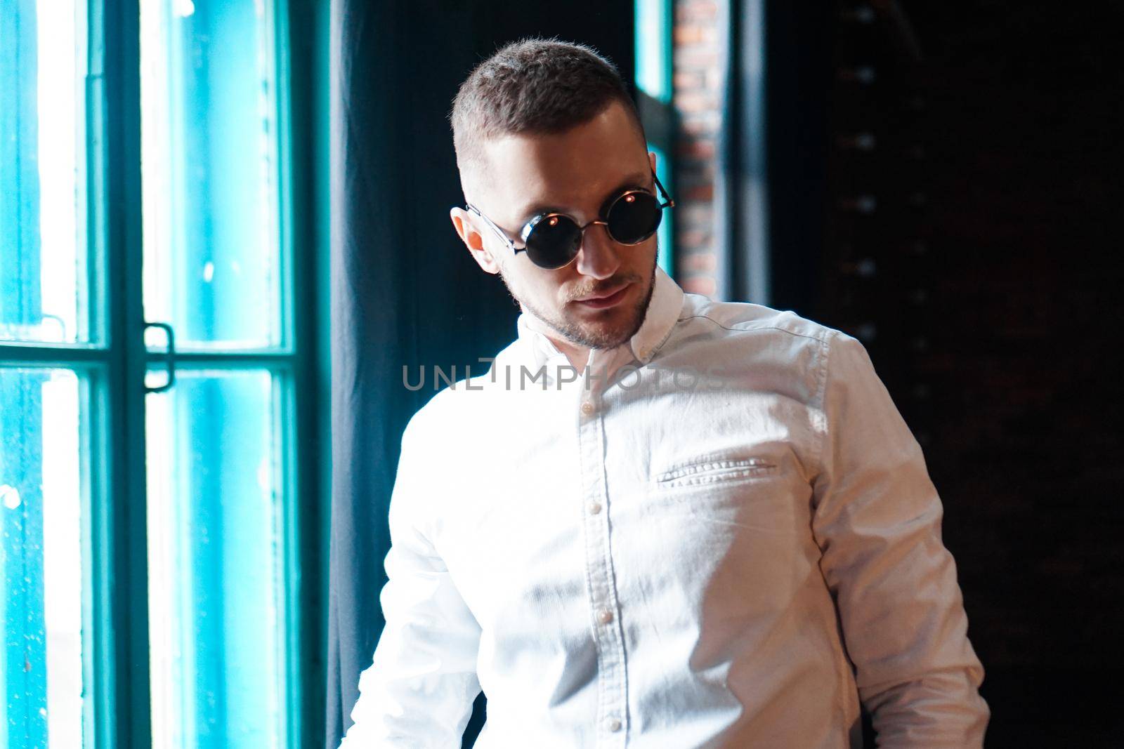 Handsome young man wearing vintage round black glasses looking forward on black background and window