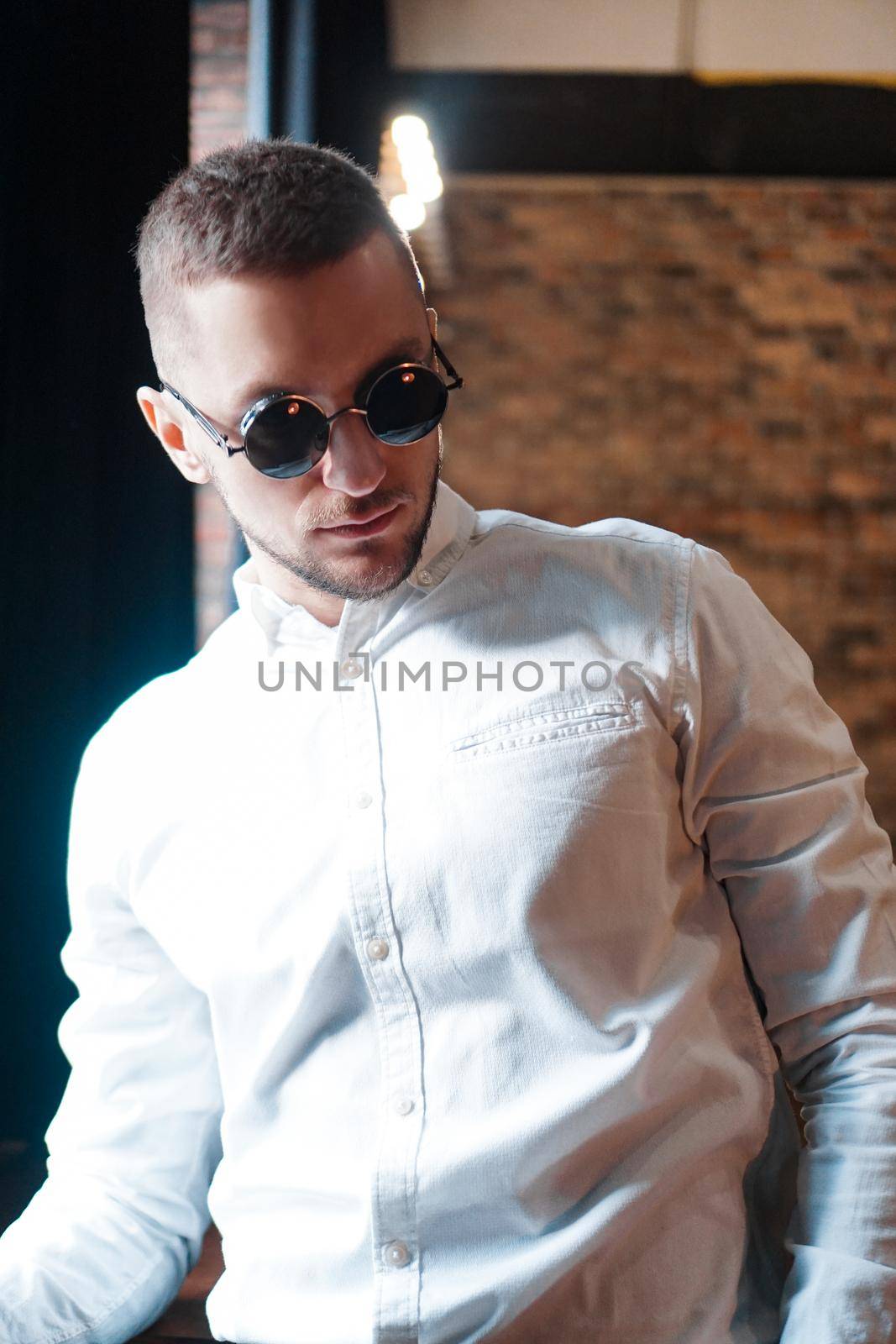 Handsome young man wearing vintage round black glasses looking forward near red brick wall