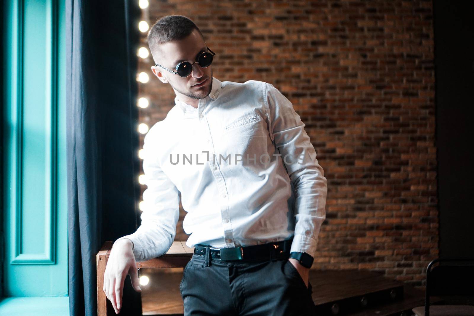 Handsome young man wearing vintage round black glasses looking forward near red brick wall