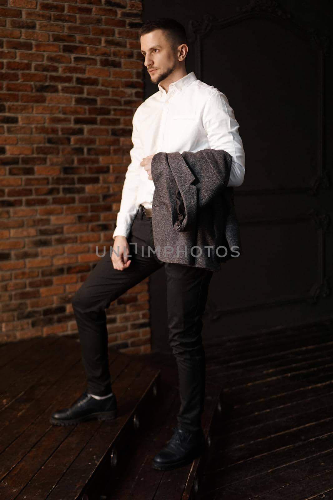Stylish young businessman in white shirt is holding a jacket, standing against brick wall - Vertical photo