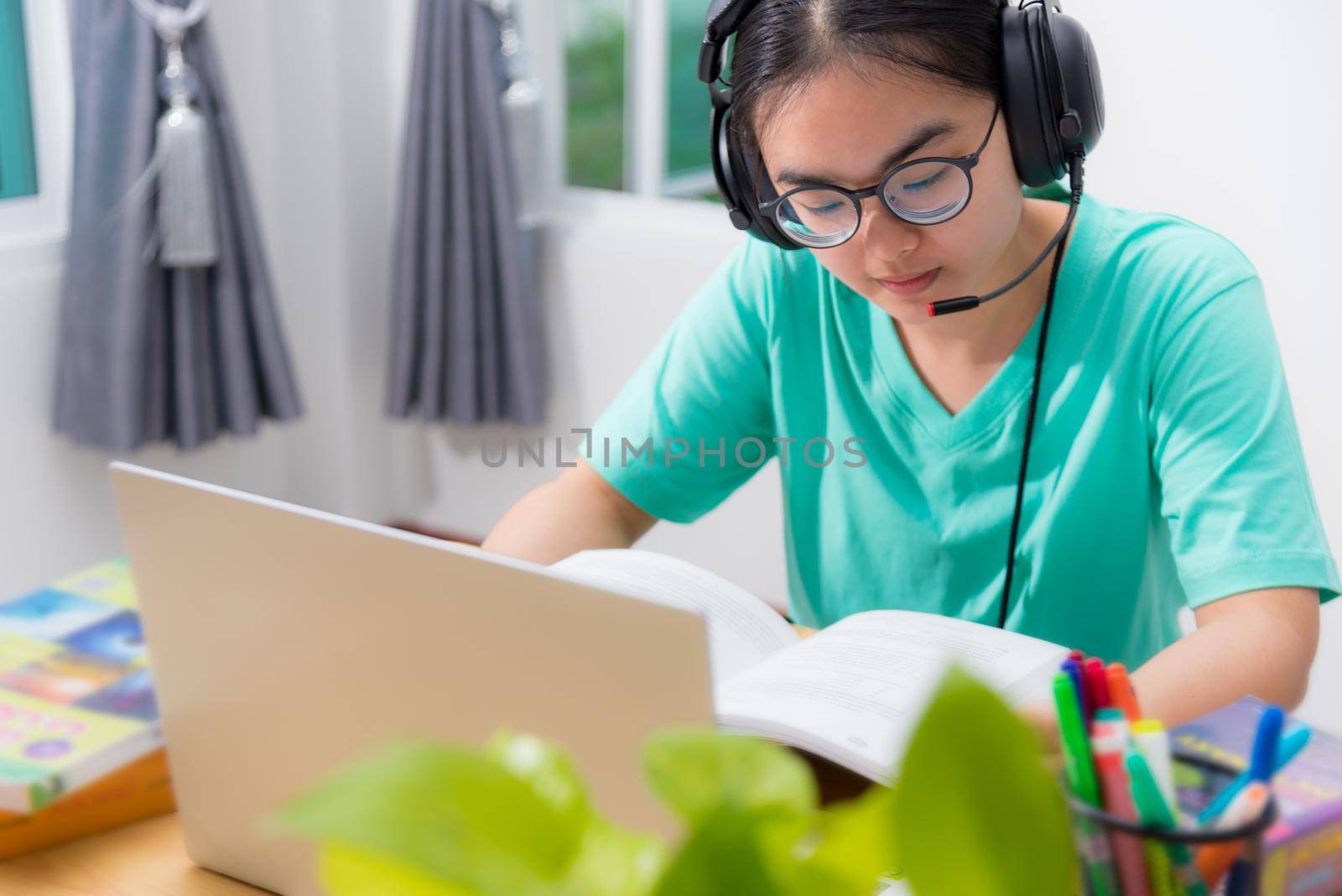 Asian woman student teenage girl with glasses headphones sitting looking serious reading a book worry using laptop computer on table learning online study. Education from a class of university at home