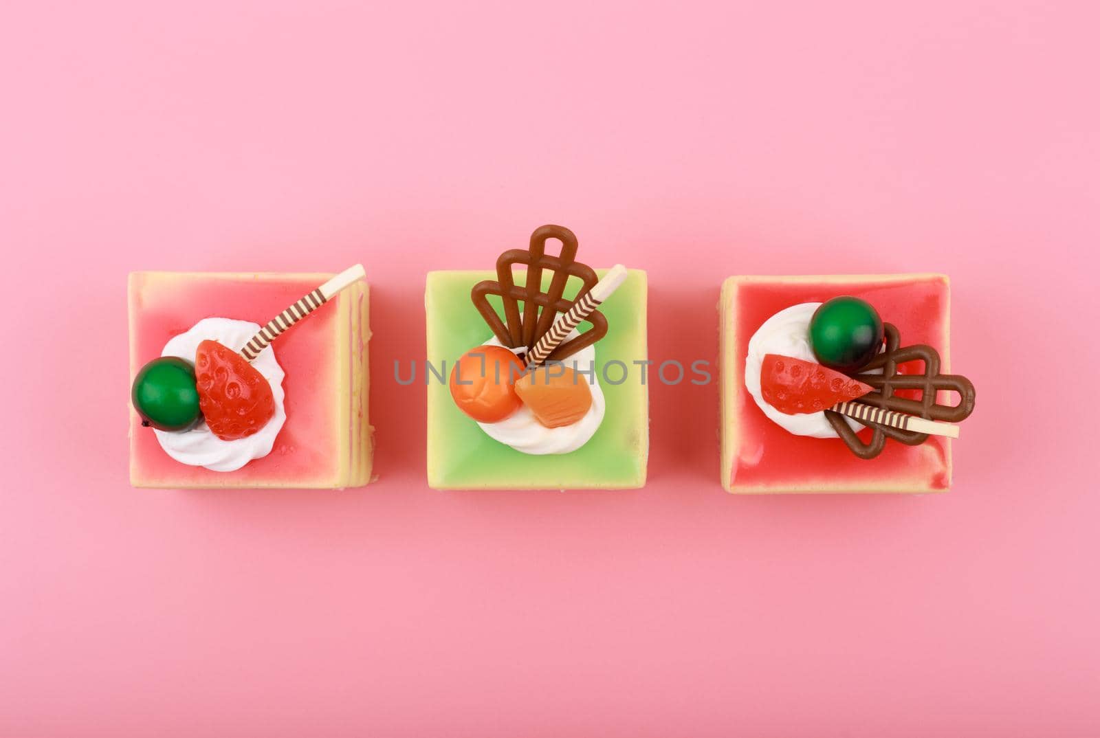 Top view of three colorful mini cakes with berries and chocolate sticks on bright pink background. Concept of sweets or cheat meal