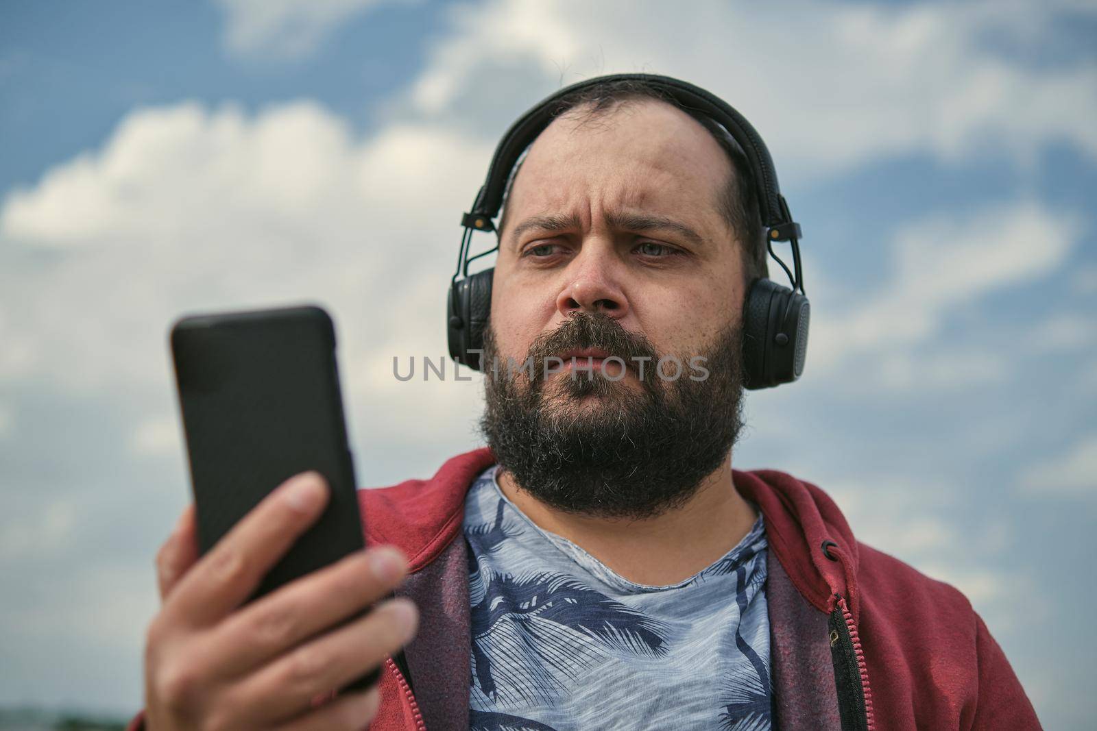 Middle-aged European man in headphones outdoors listening to music against the background of the sky, mobile phone in his hand