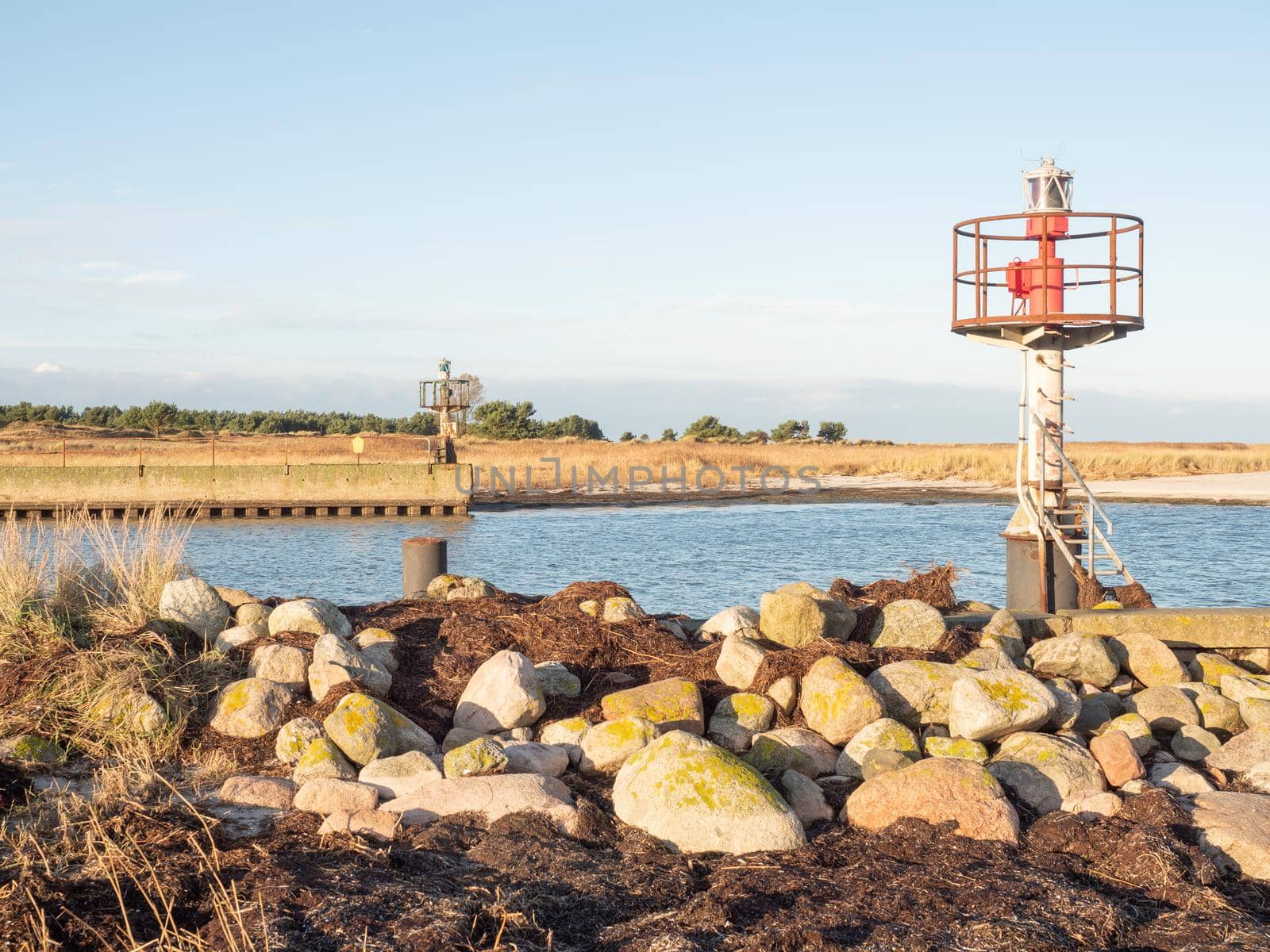 Red and white small technical lighthouse on the stony pier viewed from low angle with dry grass and white stones in foreground