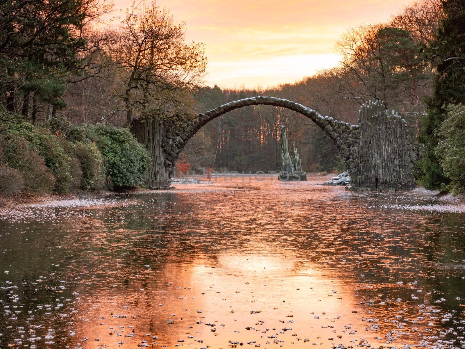 Chill autumn morning in Azalea and Rhododendron Park Kromlau. Round Arch basalt bridge over frozen lake. 