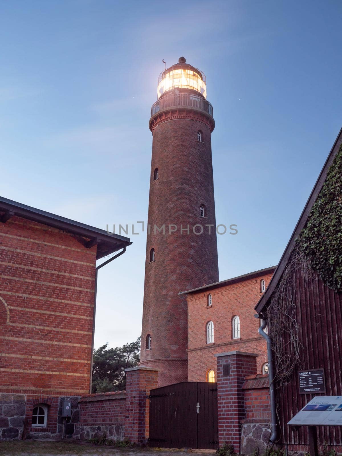 Shinning lighthouse in evening. Tower in park at Prerow built from red bricks. Tower illuminated with strong warning light dark sky in background.