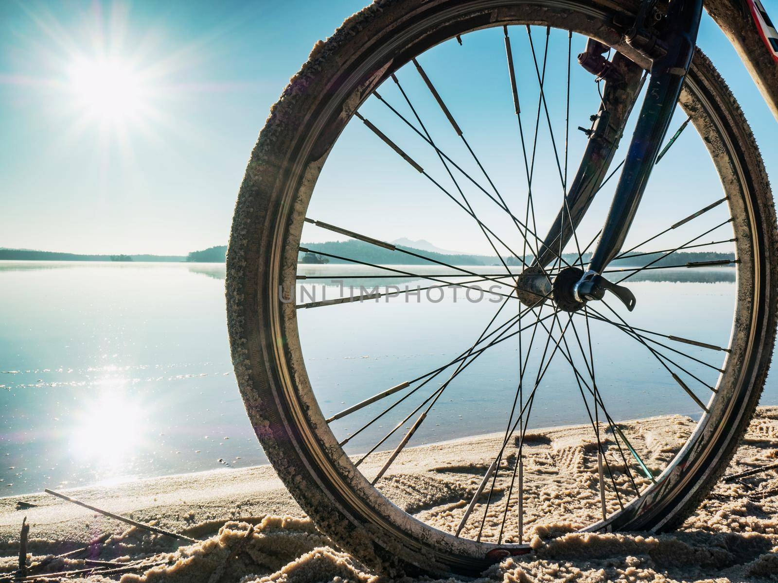 Front wheel of graviel bike in sand of beach. The sun is mirroring in water by rdonar2