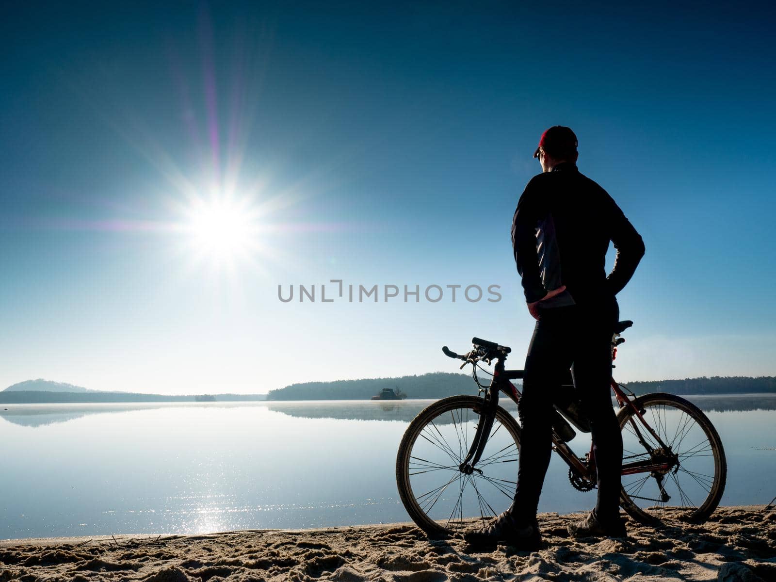 Young man staying on the shore of the lake with bicycle at sunset. Man stay at gravelbike on beach